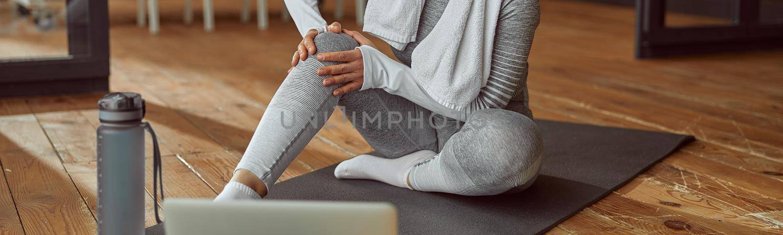Female is sitting on mat and touching joint while suffering pain during video workout on laptop at home