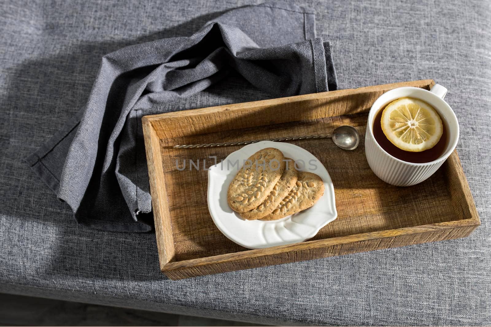 A white cup of tea with lemon, a long cupronickel spoon with a twisted handle and a saucer with three oatmeal cookies for breakfast on a wooden tray, a rag napkin on a gray sofa. Serving