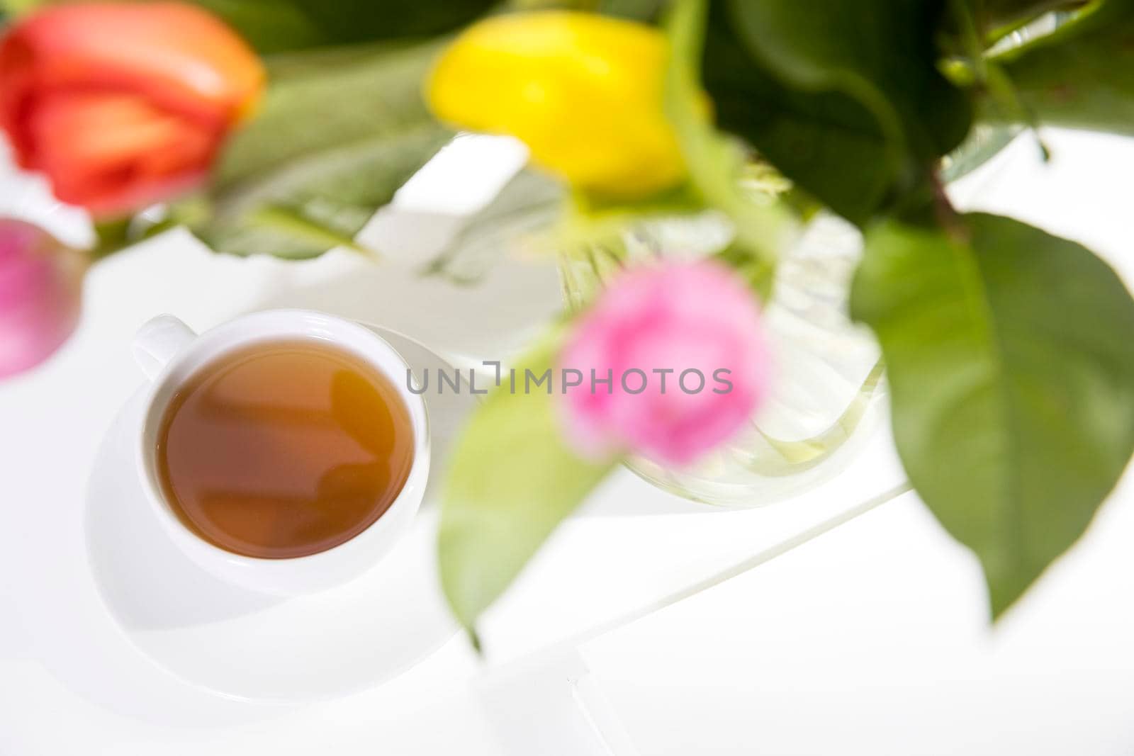 A bouquet of multi-colored tulips in a transparent vase and cup of tea and tea pot on the white table.