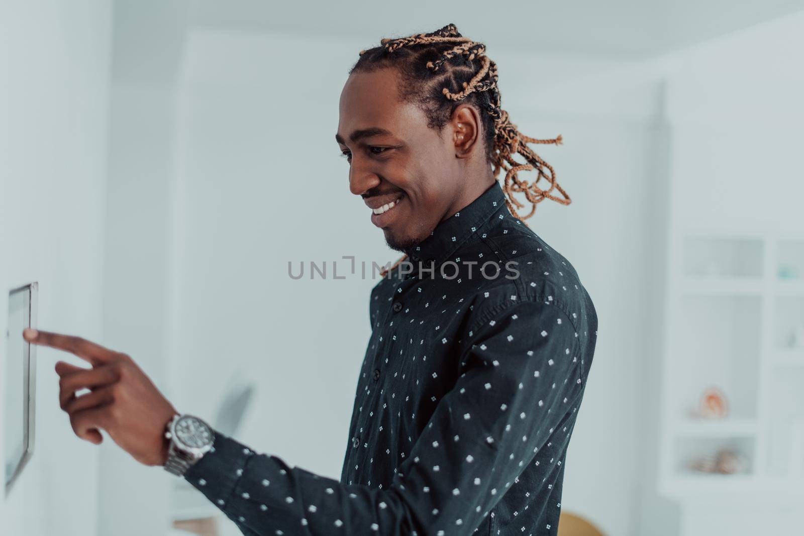 Smiling African American man using modern smart home system, controller on wall, positive young man switching temperature on thermostat or activating security alarm in apartment by dotshock