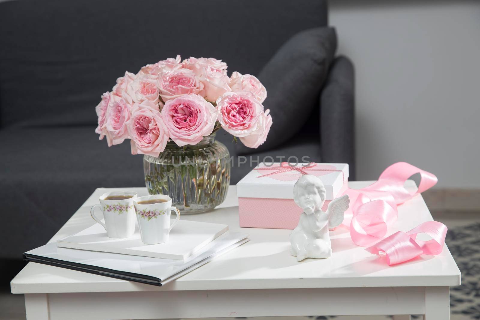 a bouquet of pink roses in a glass vase, two porcelain cups with tea, a white box with a heart with a gift, a satin ribbon, a figurine of an angel - gifts for Valentine's Day on a coffee table. by elenarostunova