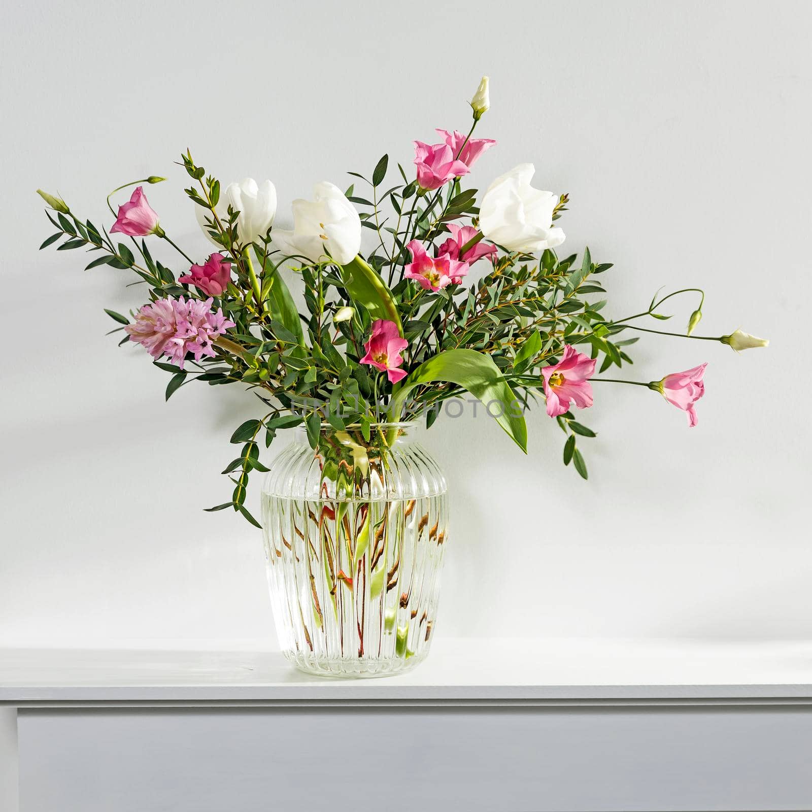 A minimalistic bouquet of white tulips, pink eustoma, hyacinth, eucalyptus in a fluted glass vase on a white panel of an artificial fireplace. by elenarostunova