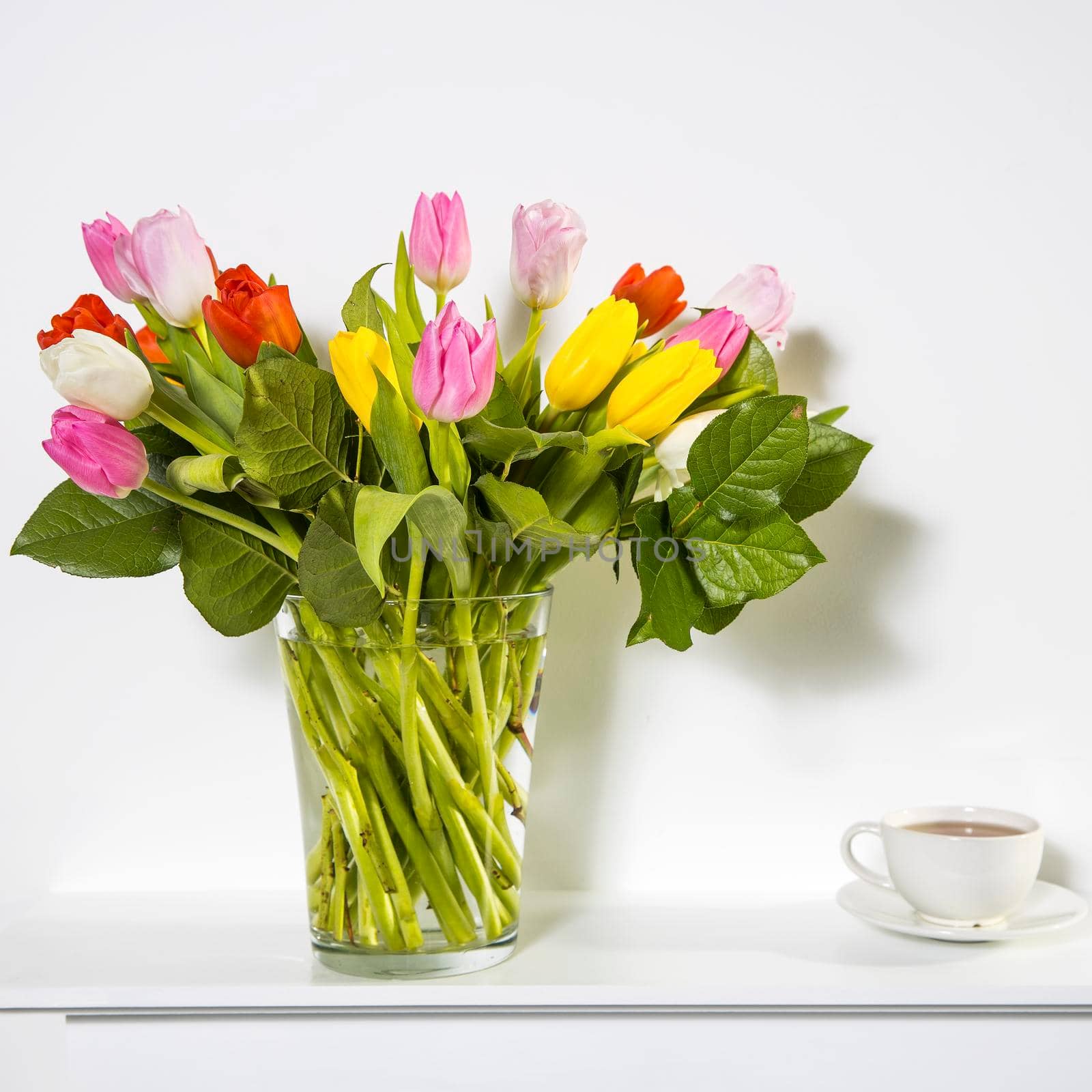 A bouquet of multi-colored tulips in a transparent vase and cup of tea on the white window.