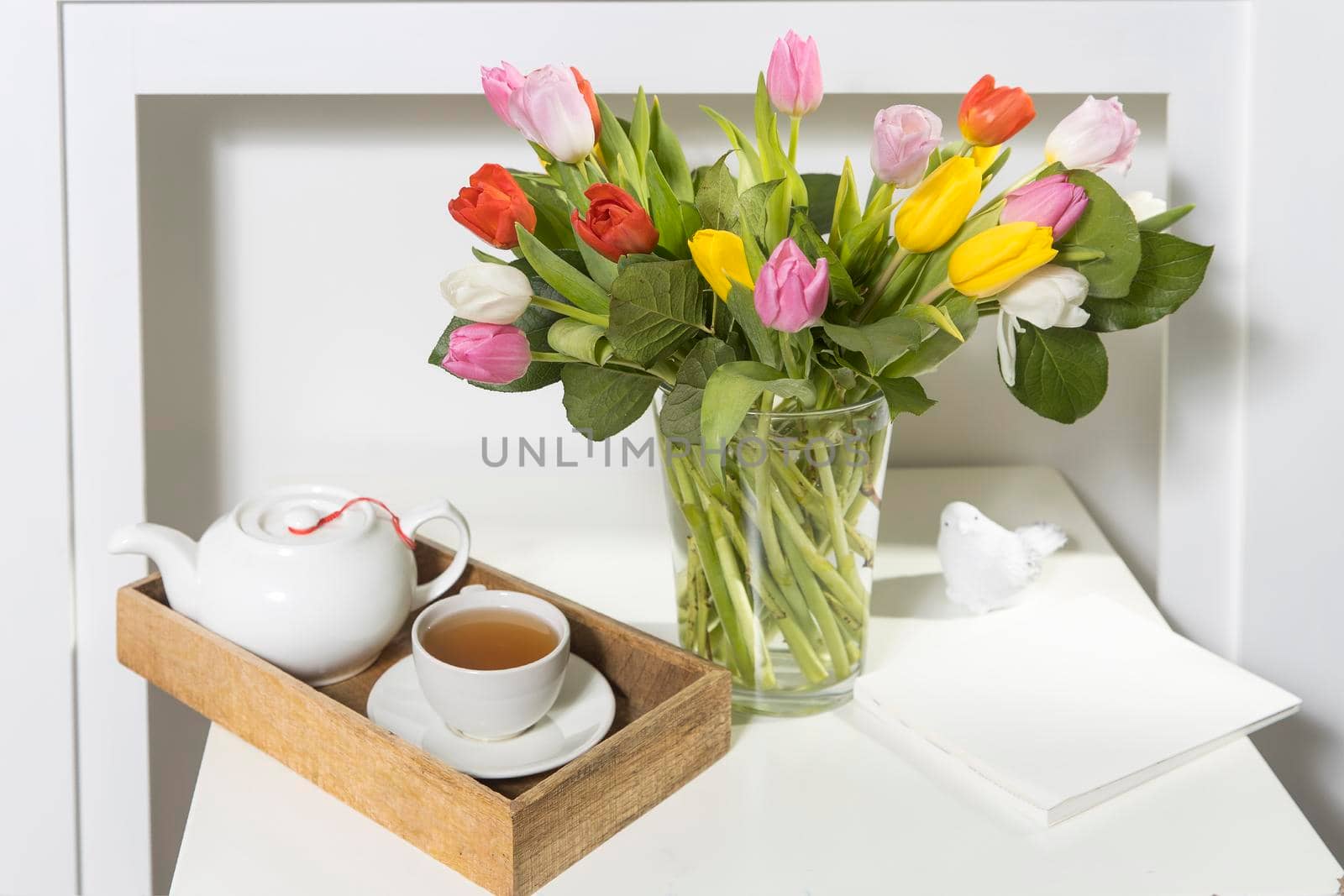 A bouquet of multi-colored tulips in a transparent vase and cup of tea on the white table. The tray is with cup of tea and tea pot.