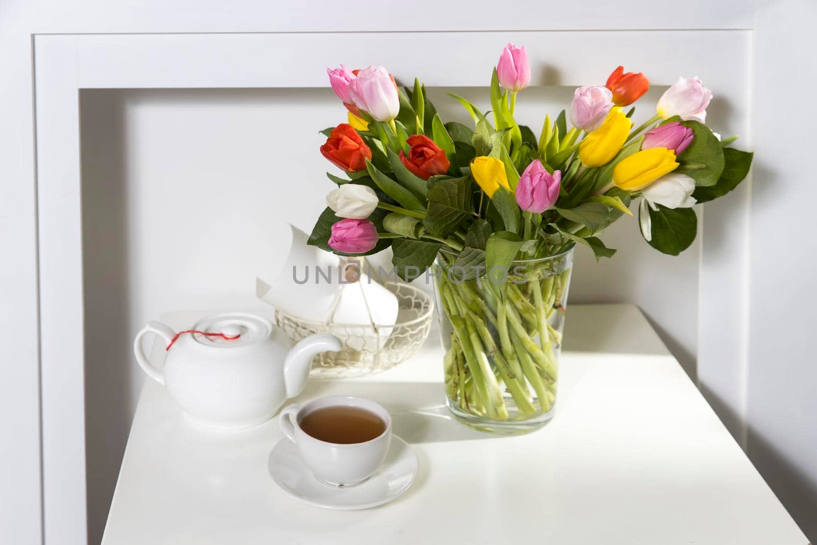 A bouquet of multi-colored tulips in a transparent vase and cup of tea and tea pot on the white table.