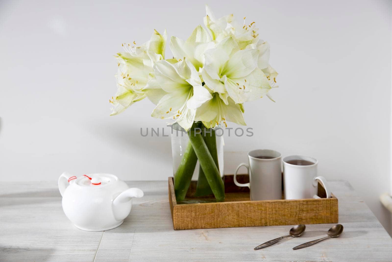 A bouquet of white lily in a glass vase on a table with two tall cups of coffee, a teapot, spoons. by elenarostunova