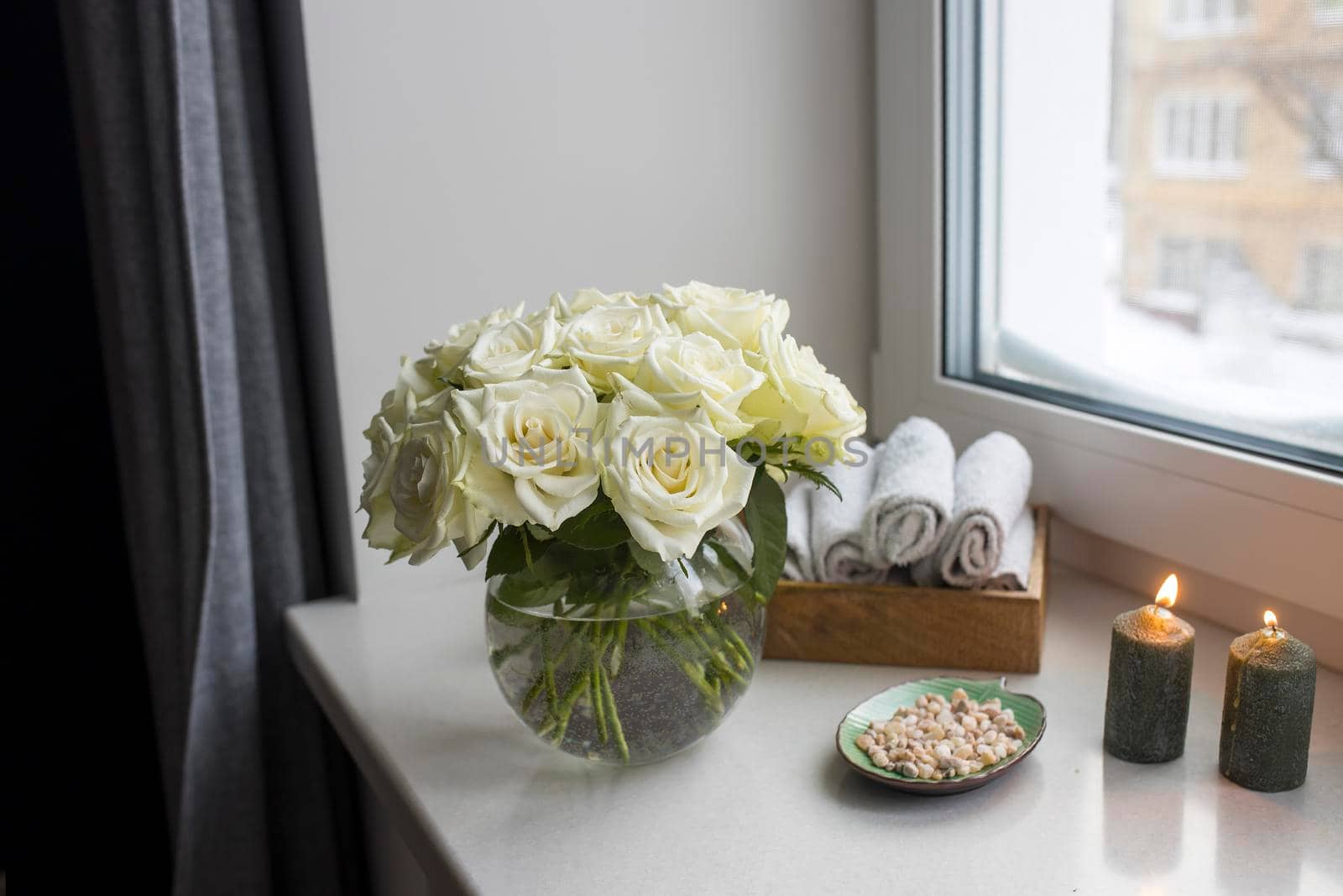 A bouquet of white roses in a round glass vase on the window with gray curtains. Tray with white small hand towels in the spa salon. by elenarostunova