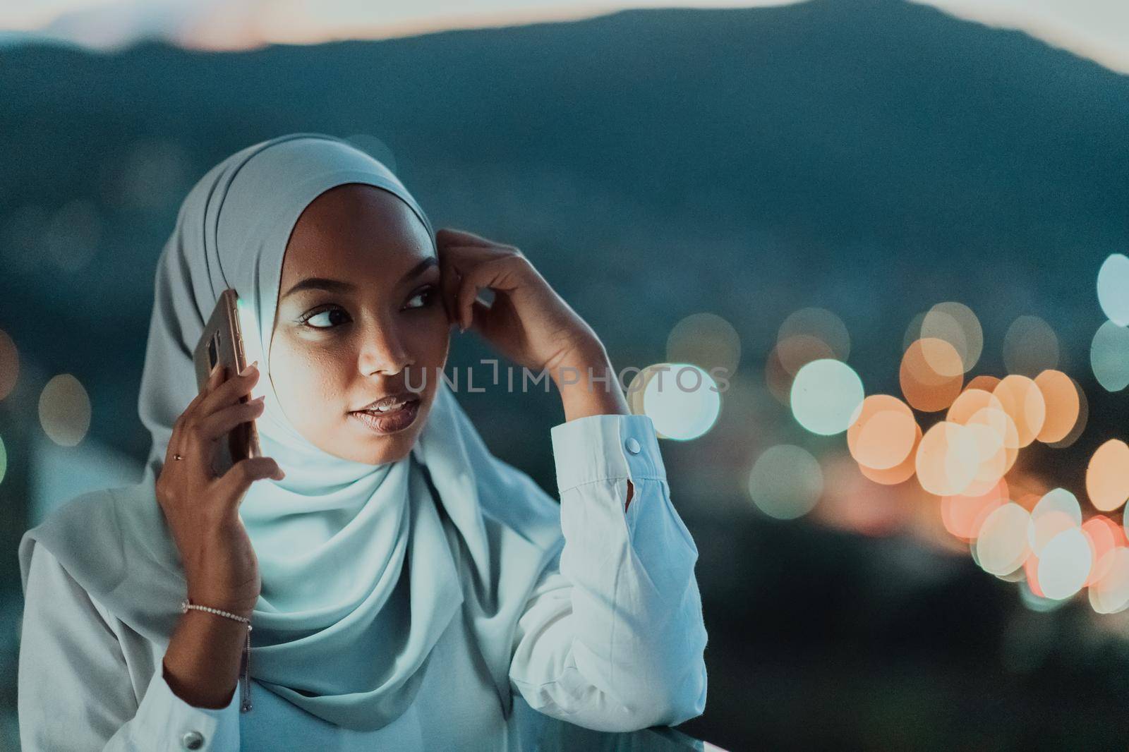 Young Muslim woman wearing scarf veil on urban city street at night texting on a smartphone with bokeh city light in the background. High-quality photo