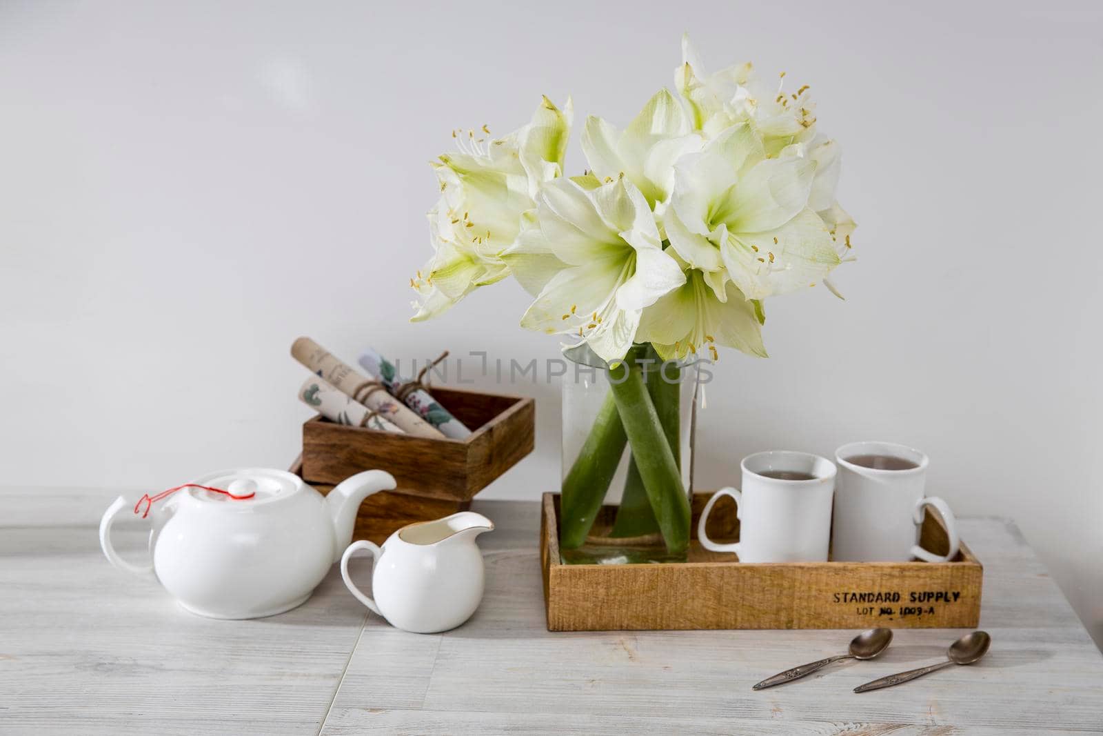 A bouquet of white lily in a glass vase on a table with two tall cups of coffee, a teapot, spoons, and a milk jug. Copy space