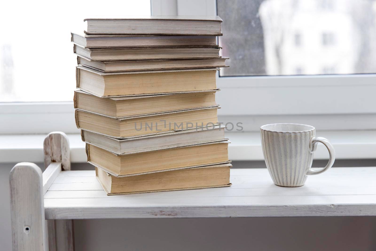 A stack of white-bound books is on the table against the background of the window. The cup of tea is next to books. School education concept. by elenarostunova