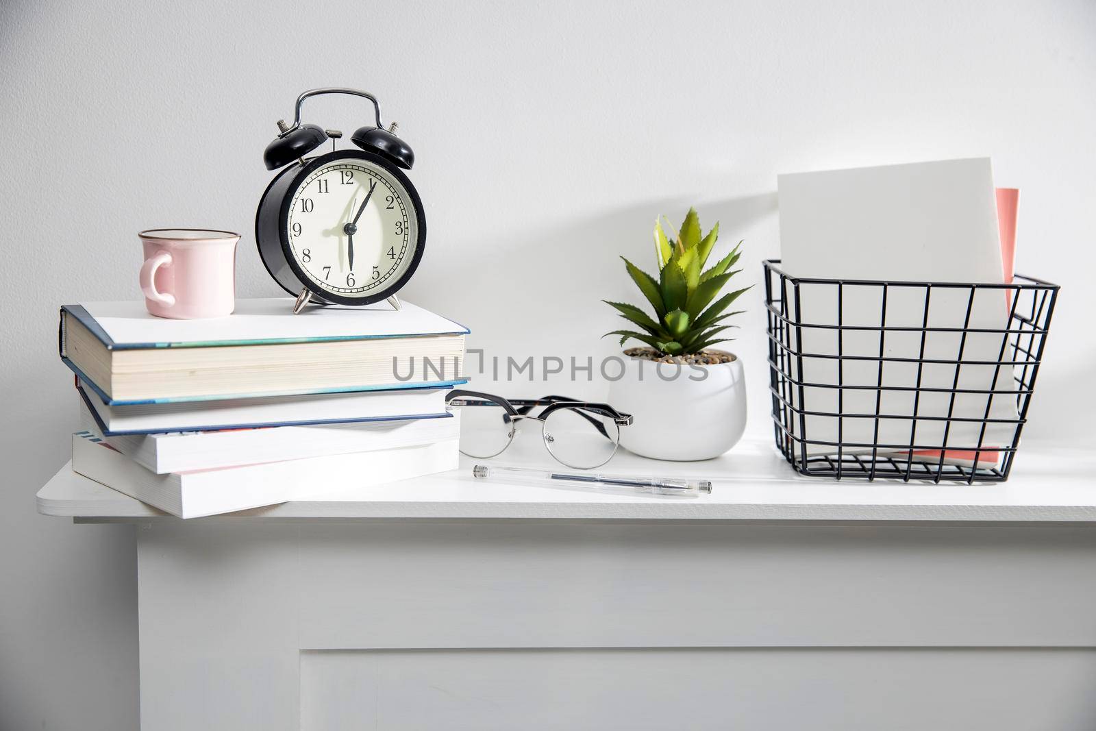 A stack of books, an artificial succulent plant in a ceramic pot, glasses, a metal basket with notebooks, a pen and a cup on a white table surface. Concept - office at home. Online training.