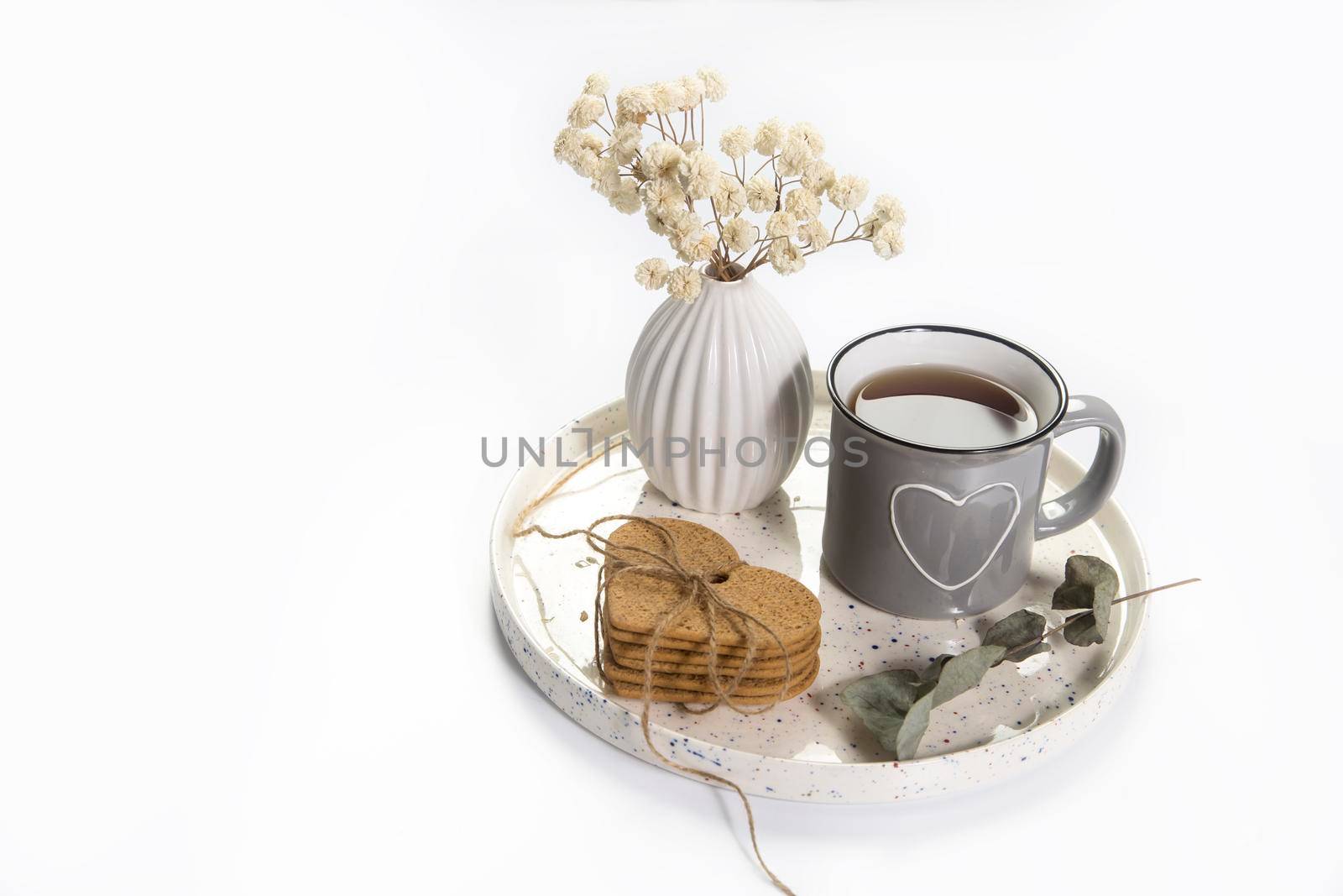 A vase with white dry flowers, a gray enamel cup with a drawn heart with tea, with gingerbread cookies stand on a tray for Valentine's Day. Copy space. Place for text. Isolated on the white background