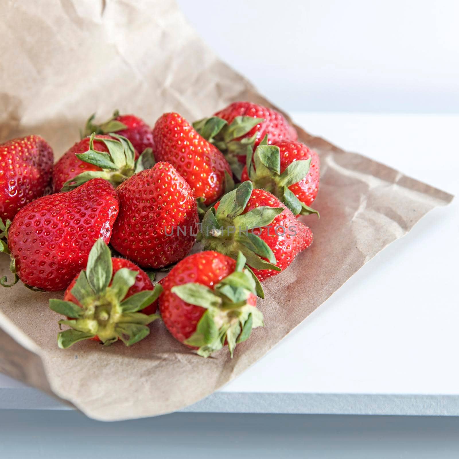 White metal basket with wooden handle with fresh strawberries on crumpled craft paper on white table. Breakfast