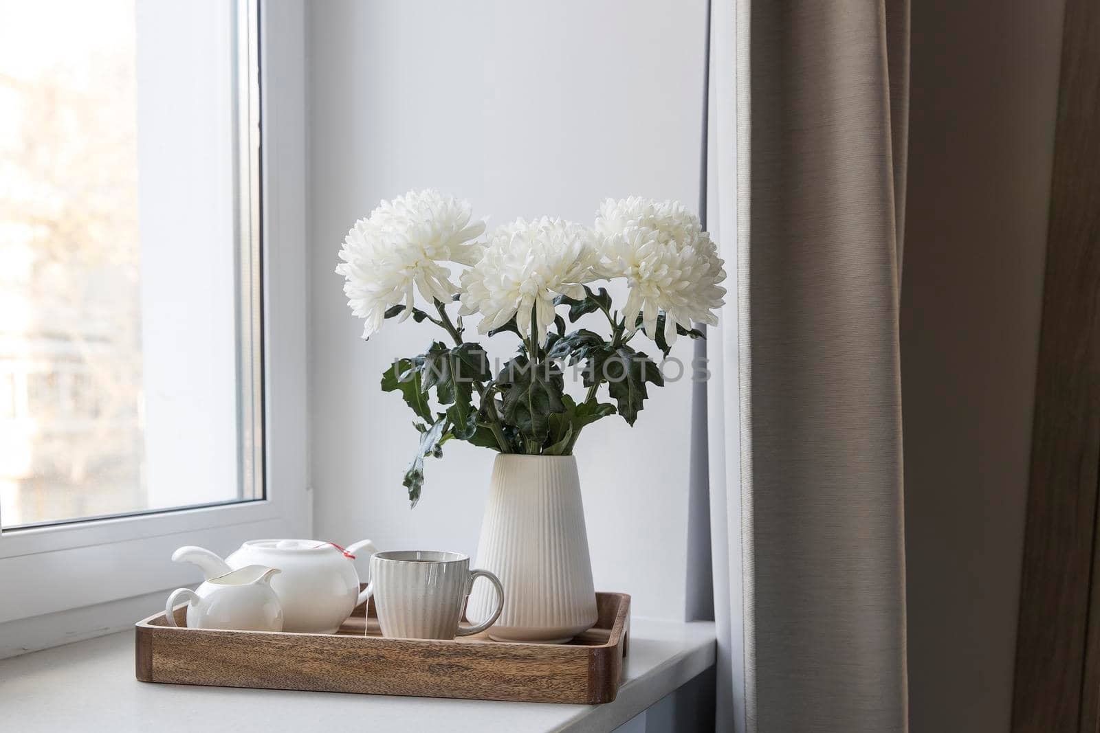 Three white chrysanthemums in a seventies-style fluted vase. Wooden tray with a kettle, milk jug and a cup of tea on the windowsill. Morning breakfast.