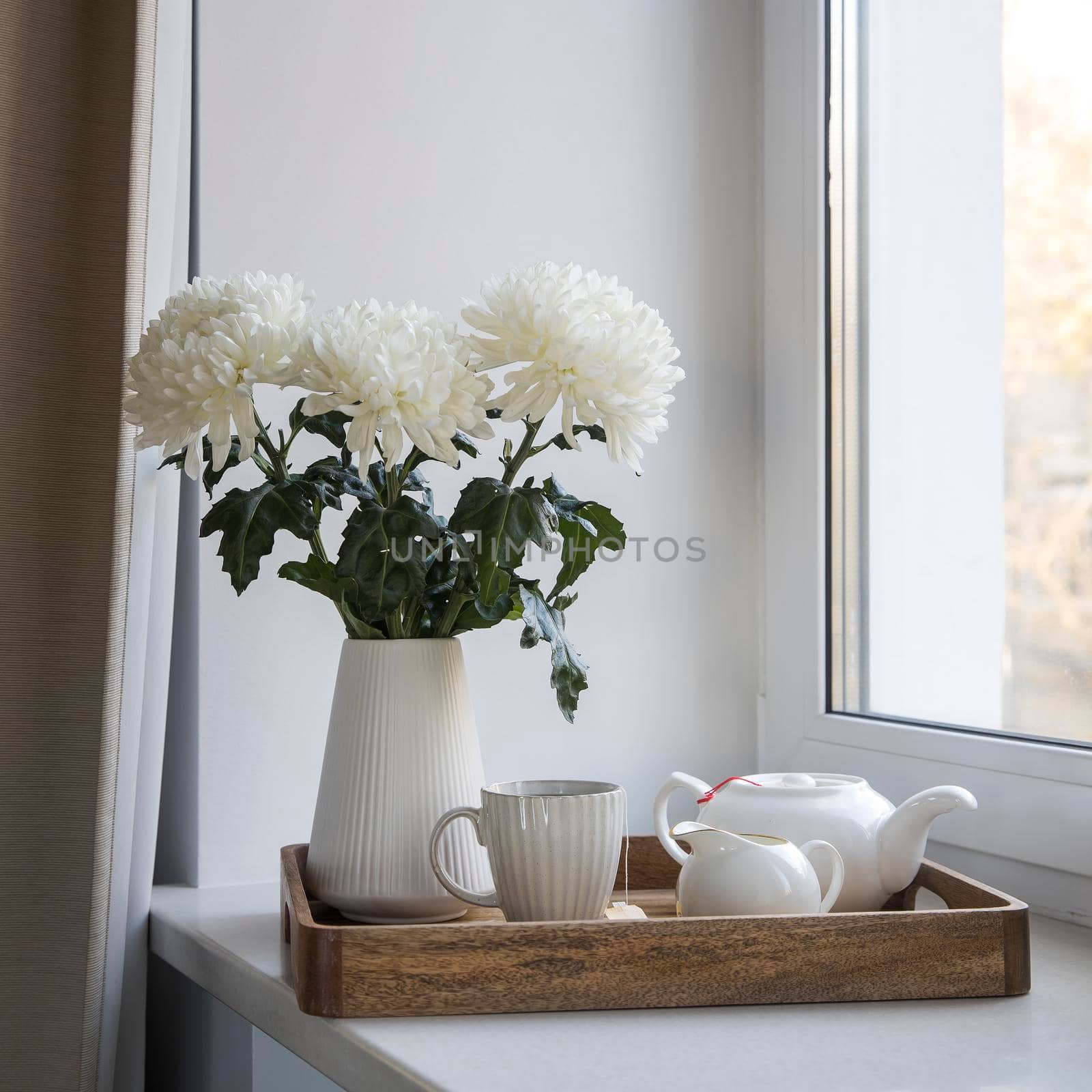 Three white chrysanthemums in a seventies-style fluted vase. Wooden tray with a kettle, milk jug and a cup of tea on the windowsill. Morning breakfast. by elenarostunova