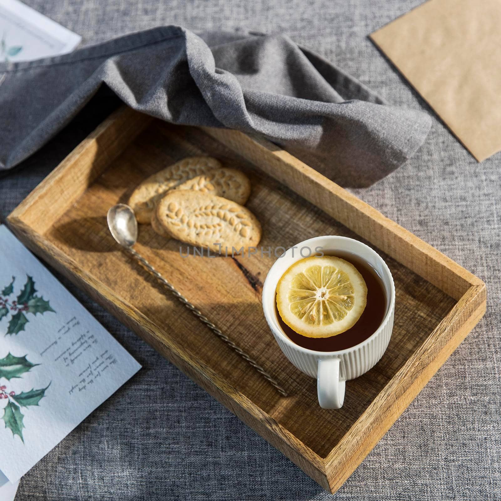 A white cup of tea with lemon, a long cupronickel spoon with a twisted handle and a saucer with three oatmeal cookies for breakfast on a wooden tray, a rag napkin on a gray sofa. by elenarostunova