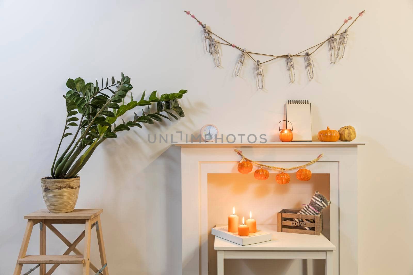 Frame with sign "Happy Halloween". Zamioculcas plant in clay pot on stool. Garland of skeletons hangs on wall. Garland of plastic pumpkins hangs from fireplace. Interior is decorated for Halloween. by elenarostunova