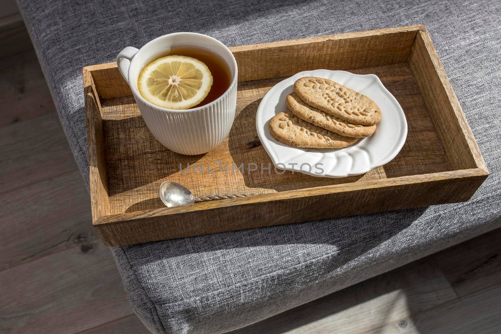 A white cup of tea with lemon, a long cupronickel spoon with a twisted handle and a saucer with three oatmeal cookies for breakfast on a wooden tray, a rag napkin on a gray sofa. by elenarostunova