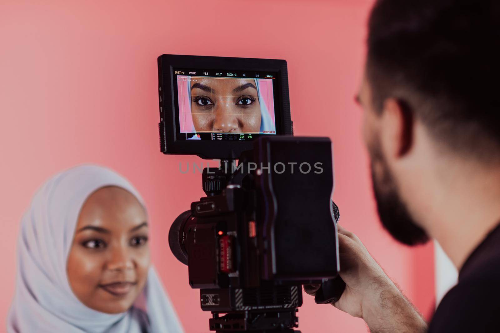 Videographer in digital studio recording video on professional camera by shooting female Muslim woman wearing hijab scarf plastic pink background. High-quality photo