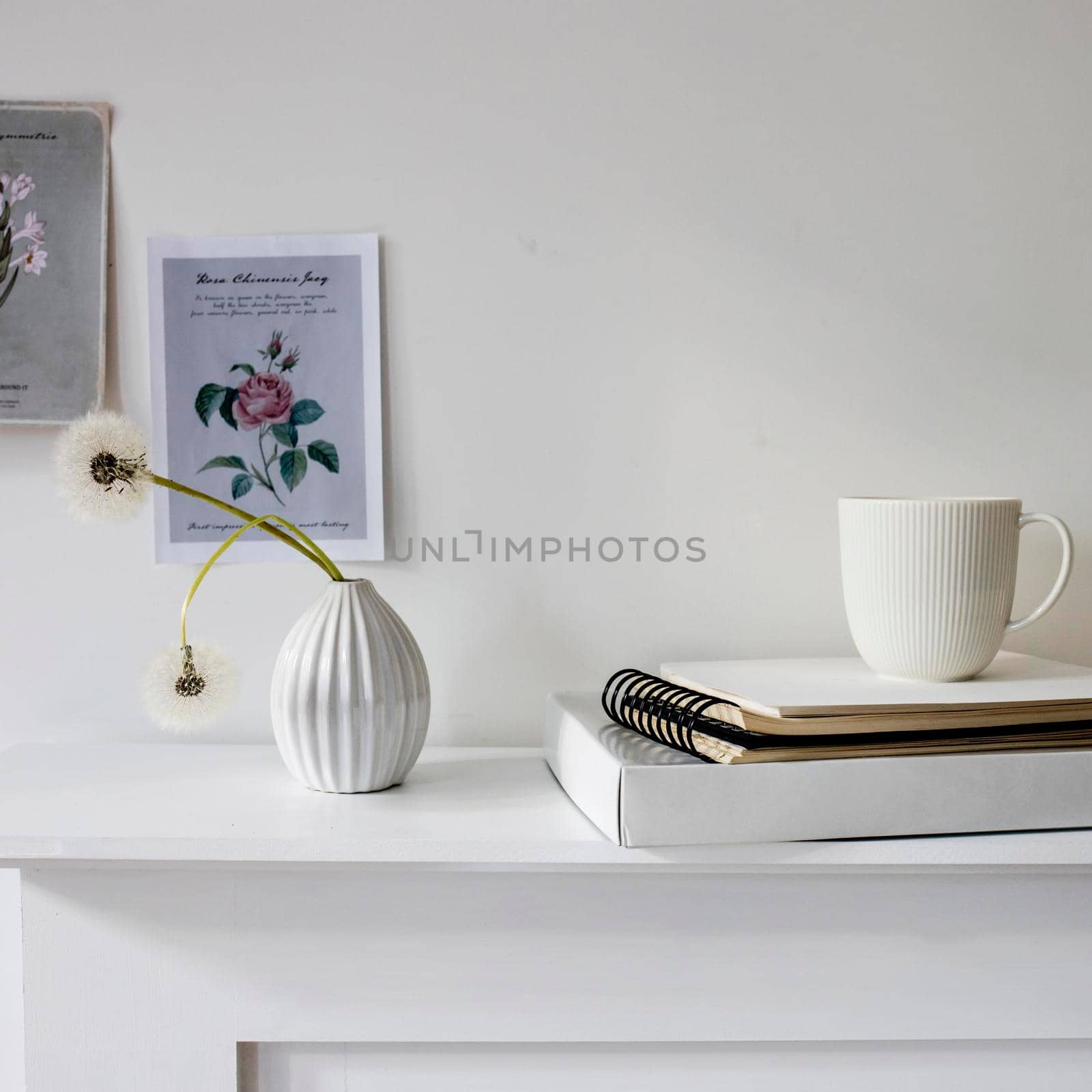 A seventies-style fluted vase with two fluffy dandelions is on the dresser. A small picture with a flower in pastel colors on the wall. A white cup of tea stands on spring-loaded notebooks. by elenarostunova