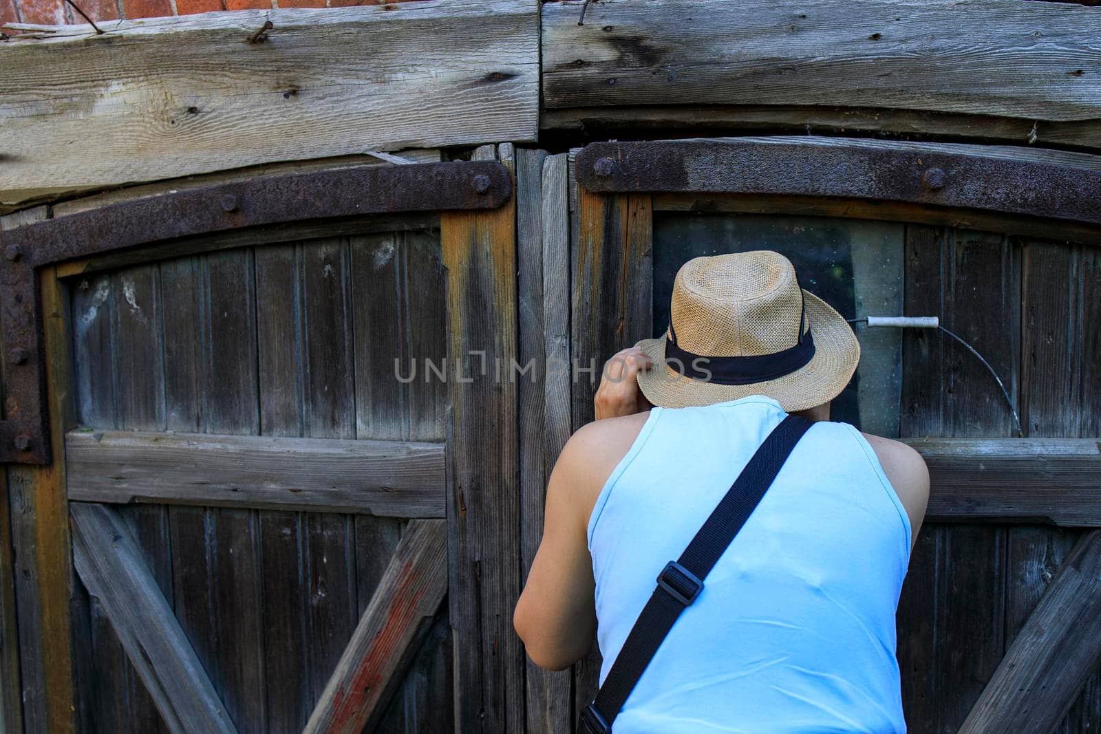 A girl in a straw hat peers through the crack in the door of an old barn by elenarostunova