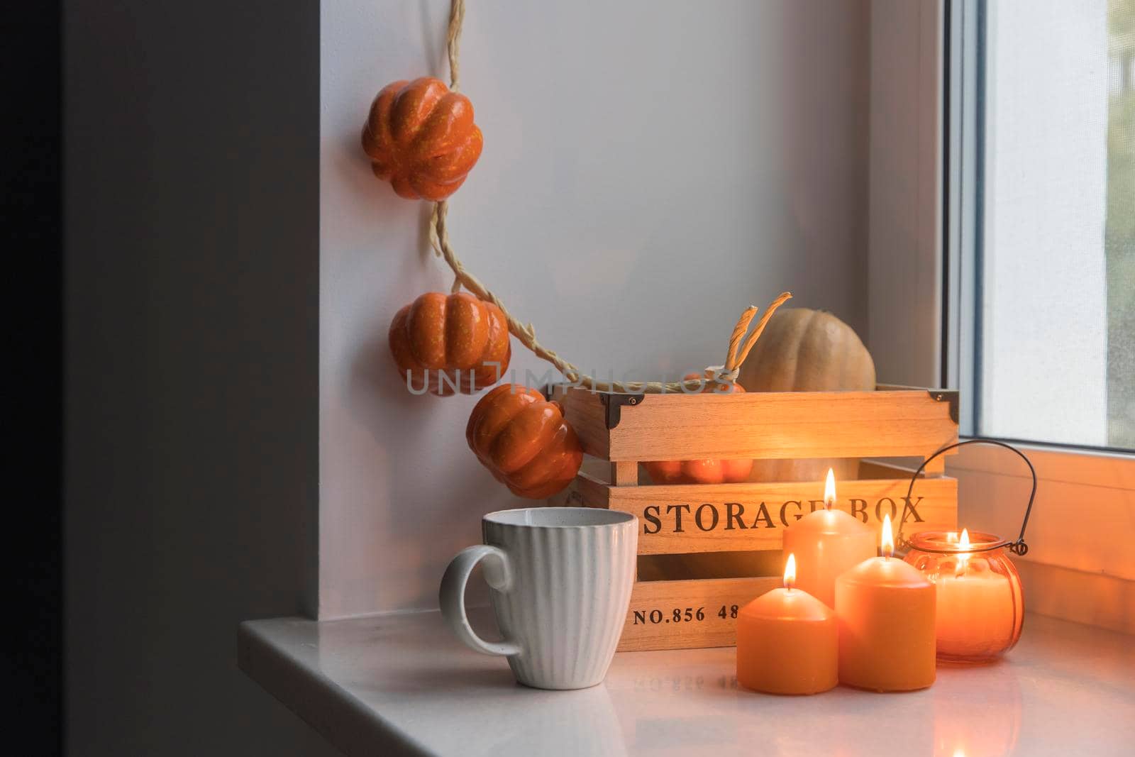 Preparing your home for Halloween. A wooden pumpkin box, a faux pumpkin garland, orange candles, a cup of tea and a candle lantern decorate the window.