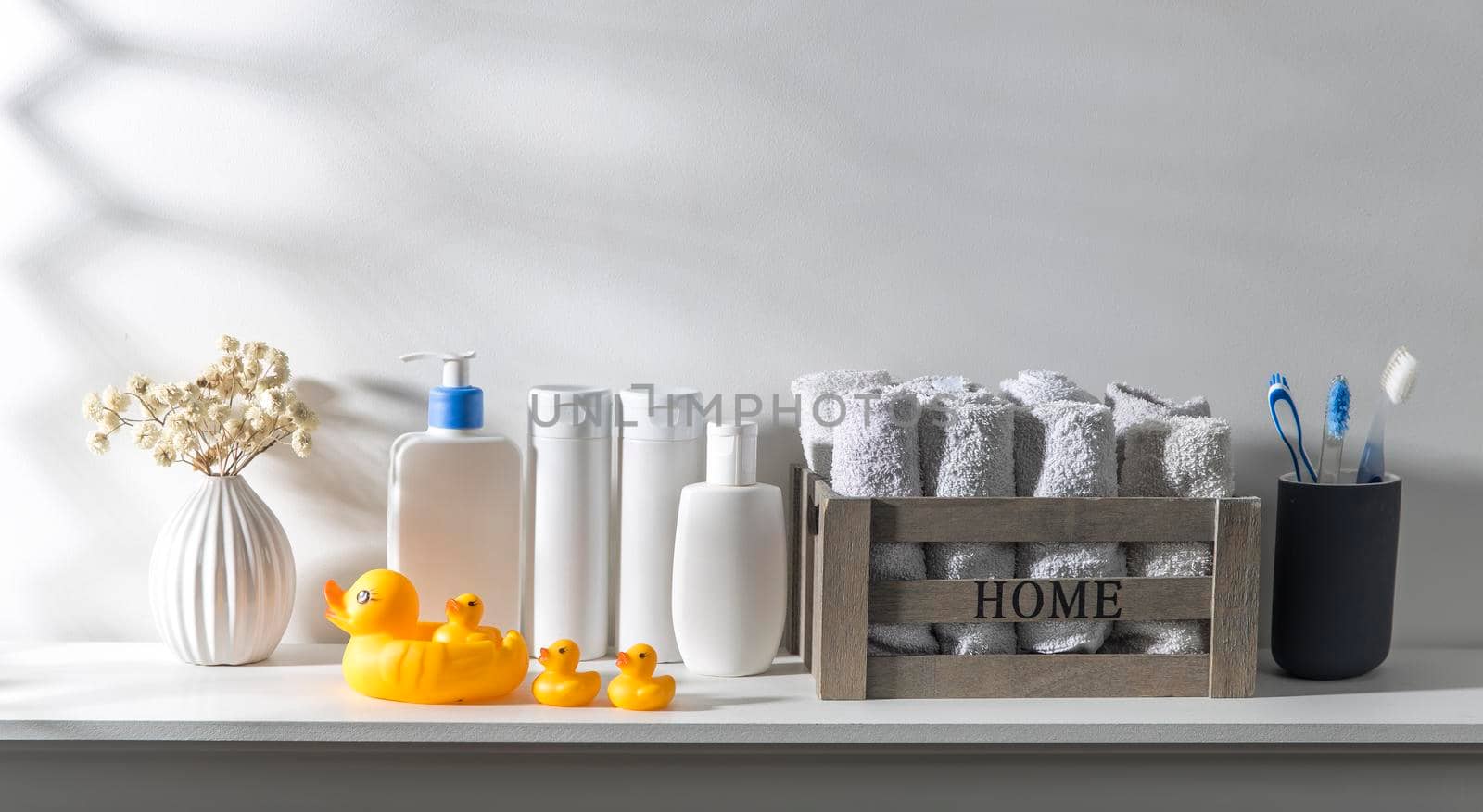 Shelf in the bathroom. Bouquet of gypsophila in white corrugated vase in style of the seventies, bottles of shampoo and cream, small face towels in container and yellow rubber ducks are on the shelf.