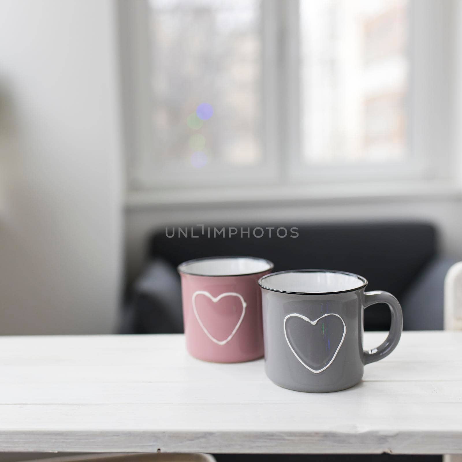 Gray and pink with hearts enamel cups sit on the table in front of the window. Valentine's Day.