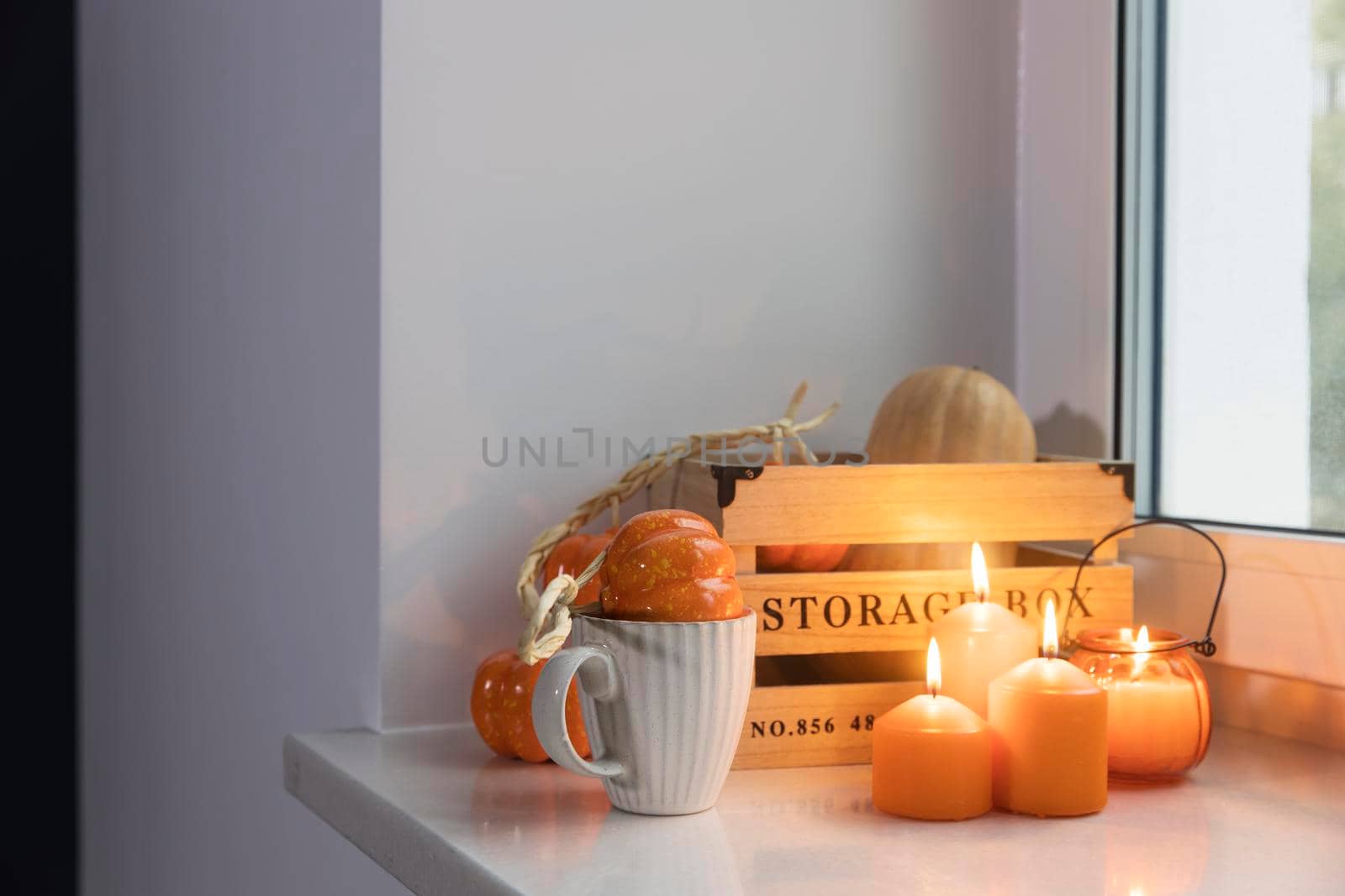 Preparing your home for Halloween. A wooden pumpkin box, faux pumpkin garland, orange candles, a cup of tea and a candle lantern decorate the window. by elenarostunova