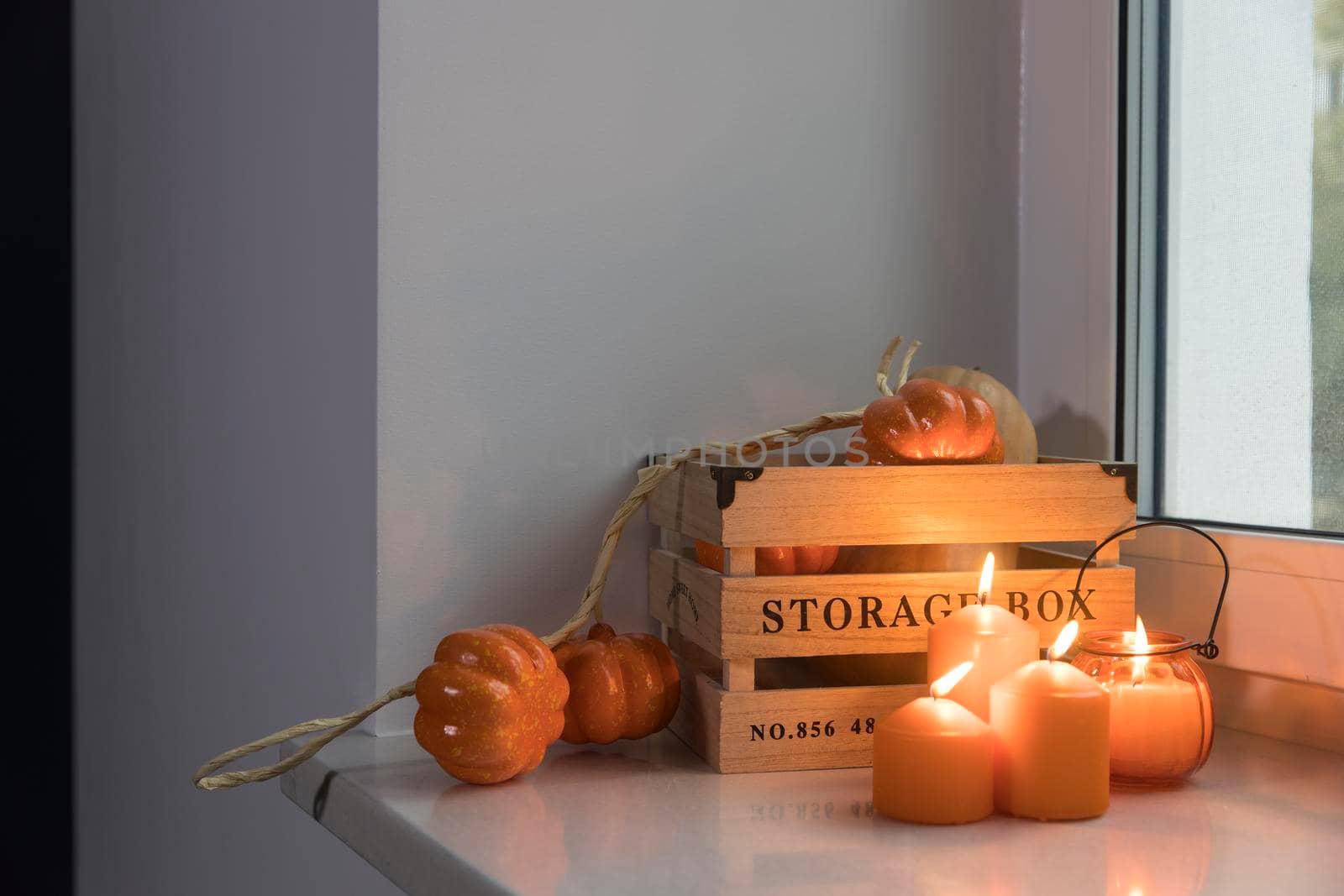 Preparing your home for Halloween. A wooden pumpkin box, a faux pumpkin garland, orange candles, a cup of tea and a candle lantern decorate the window.