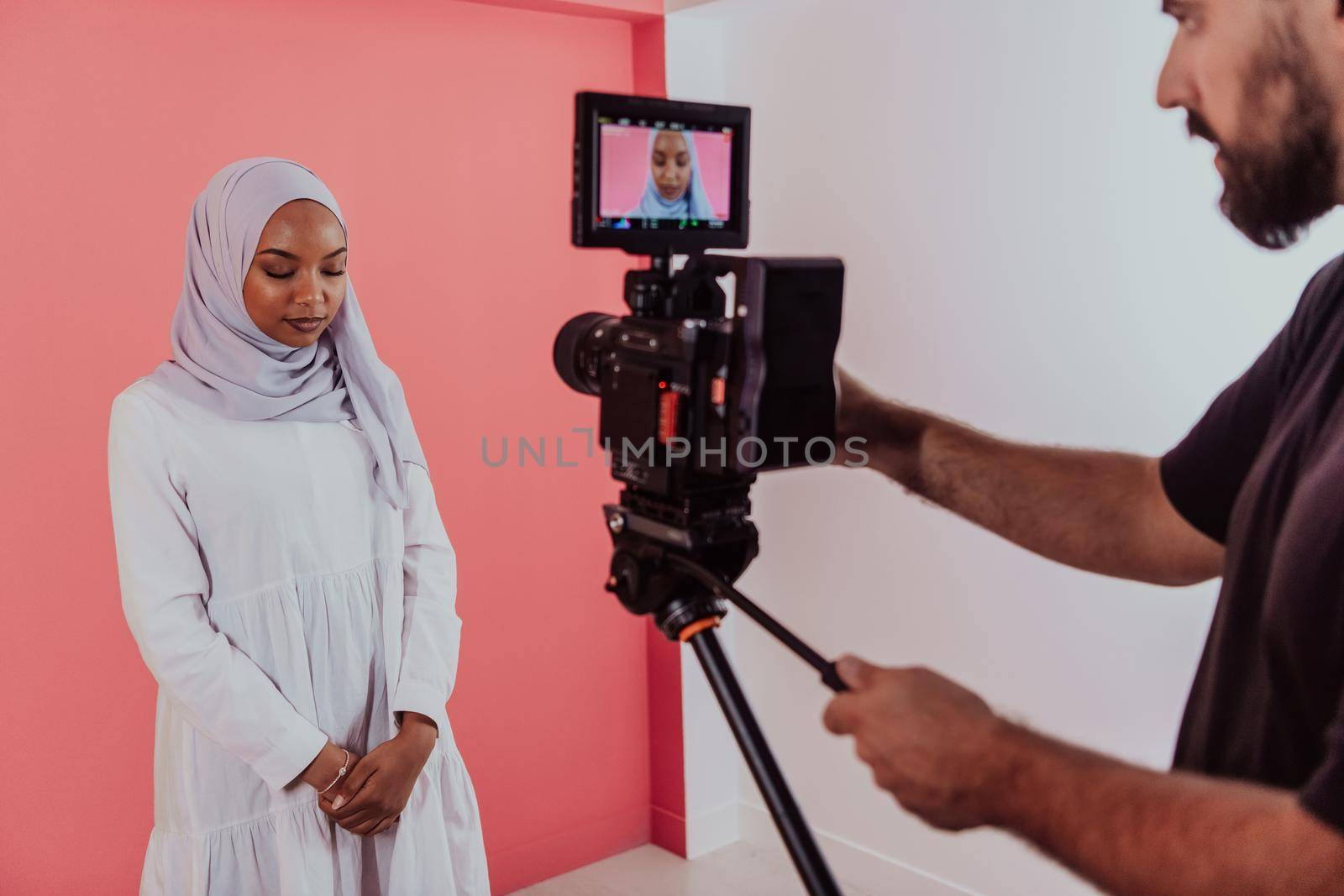 Videographer in digital studio recording video on professional camera by shooting female Muslim woman wearing hijab scarf plastic pink background. High-quality photo