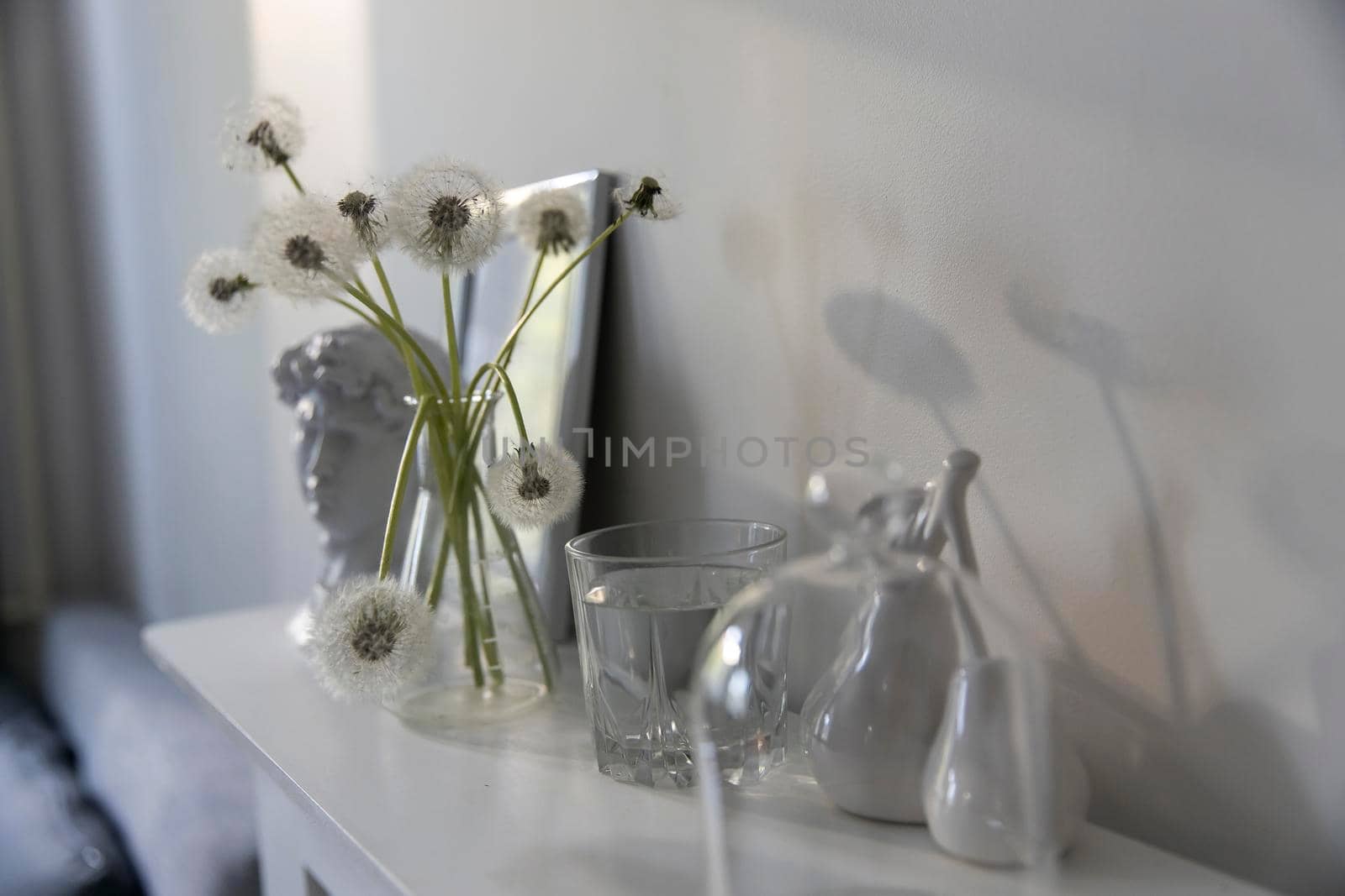 A bunch of fluffy dandelions in a chemical flask, an empty photo frame, a cup of espresso coffee, and a plaster head of David on a white chest of drawers. Copy space