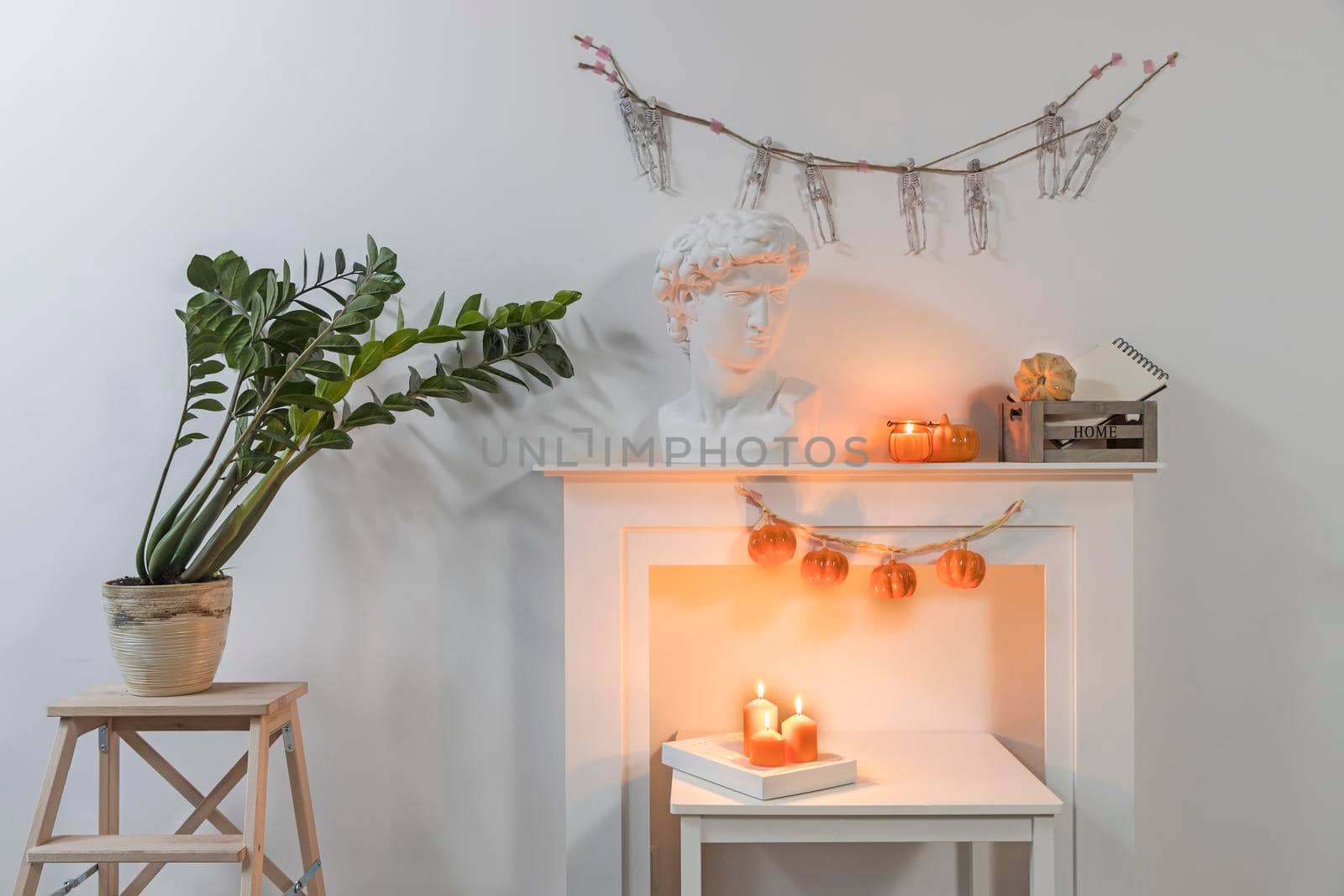 Apollo's plaster head. Zamioculcas plant in clay pot on stool. A garland of skeletons hangs on wall. A garland of plastic pumpkins hangs from fireplace. The interior is decorated for Halloween.