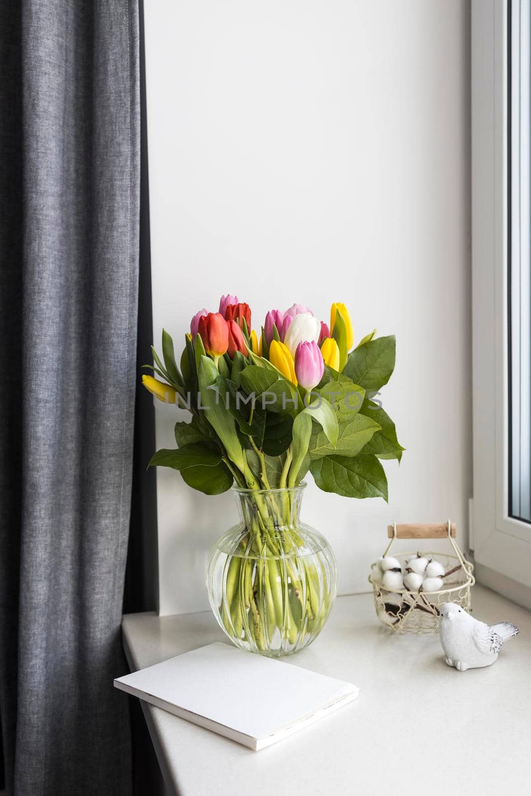 A bouquet of multi-colored tulips in a transparent vase on the windowsill on a holiday. White notebook, wicker metal basket with cotton flowers inside. Bird figurine. by elenarostunova