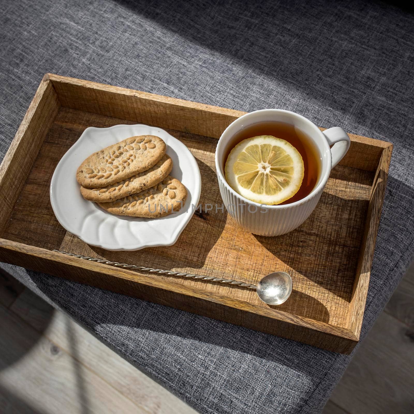 A white cup of tea with lemon, a long cupronickel spoon with a twisted handle and a saucer with three oatmeal cookies for breakfast on a wooden tray, a rag napkin on a gray sofa. by elenarostunova