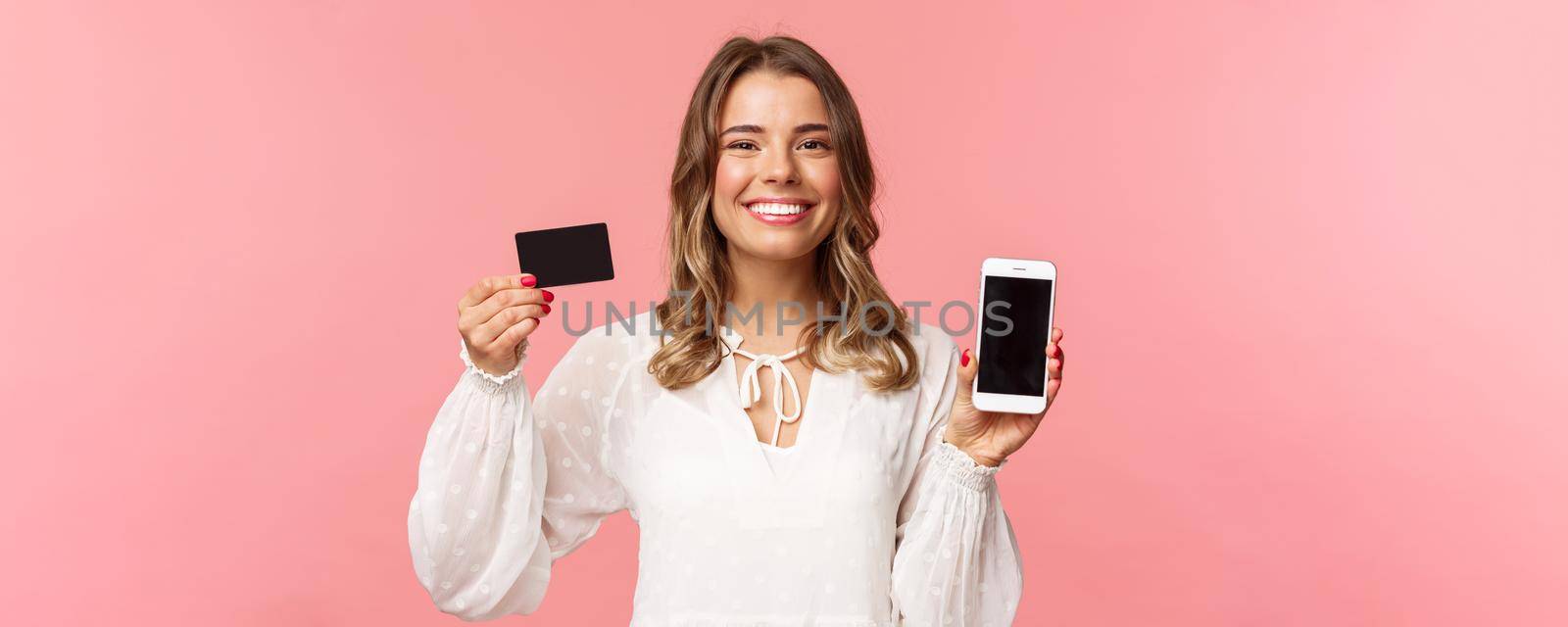 Finance, shopping and technology concept. Close-up portrait of cheerful, romantic blond cute girl in white dress, holding credit card and mobile phone, showing smartphone display, application promo.