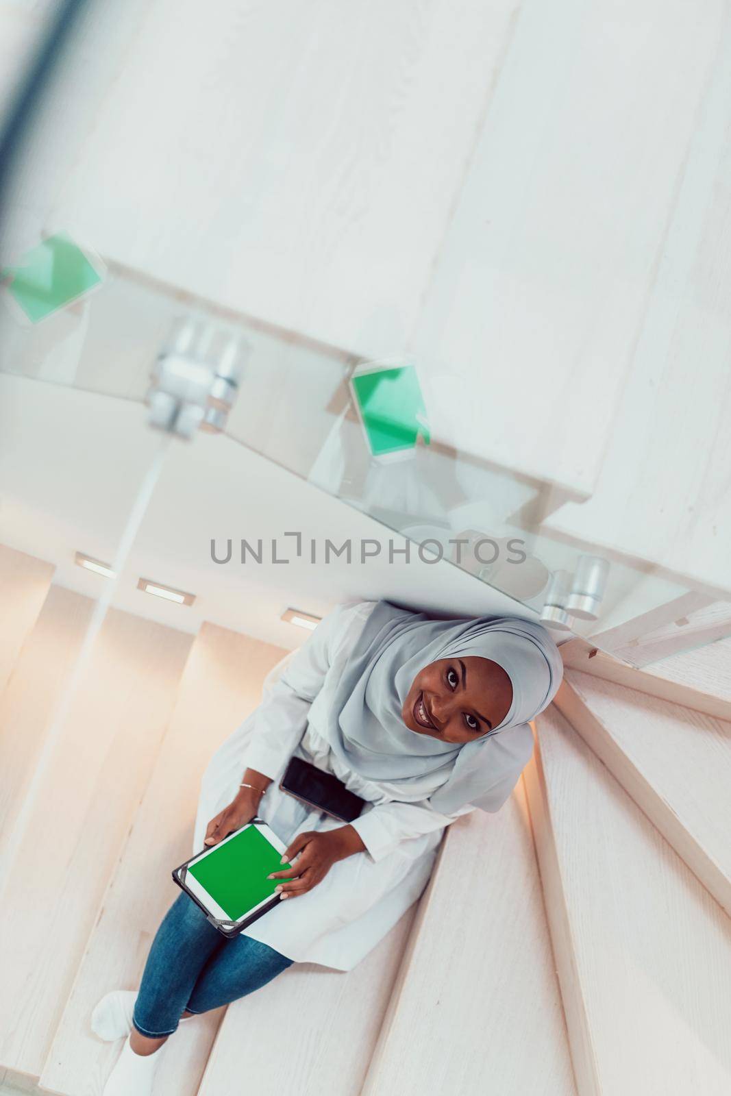 Young african modern muslim woman using tablet computer while sitting on the stairs at home wearing hijab clothes top view by dotshock