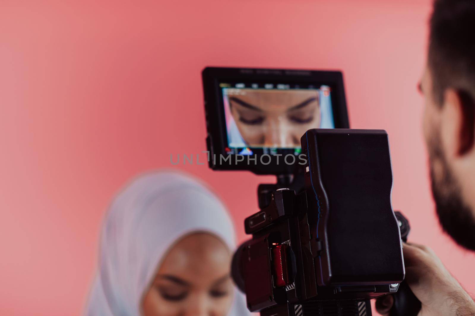 Videographer in digital studio recording video on professional camera by shooting female Muslim woman wearing hijab scarf plastic pink background. by dotshock