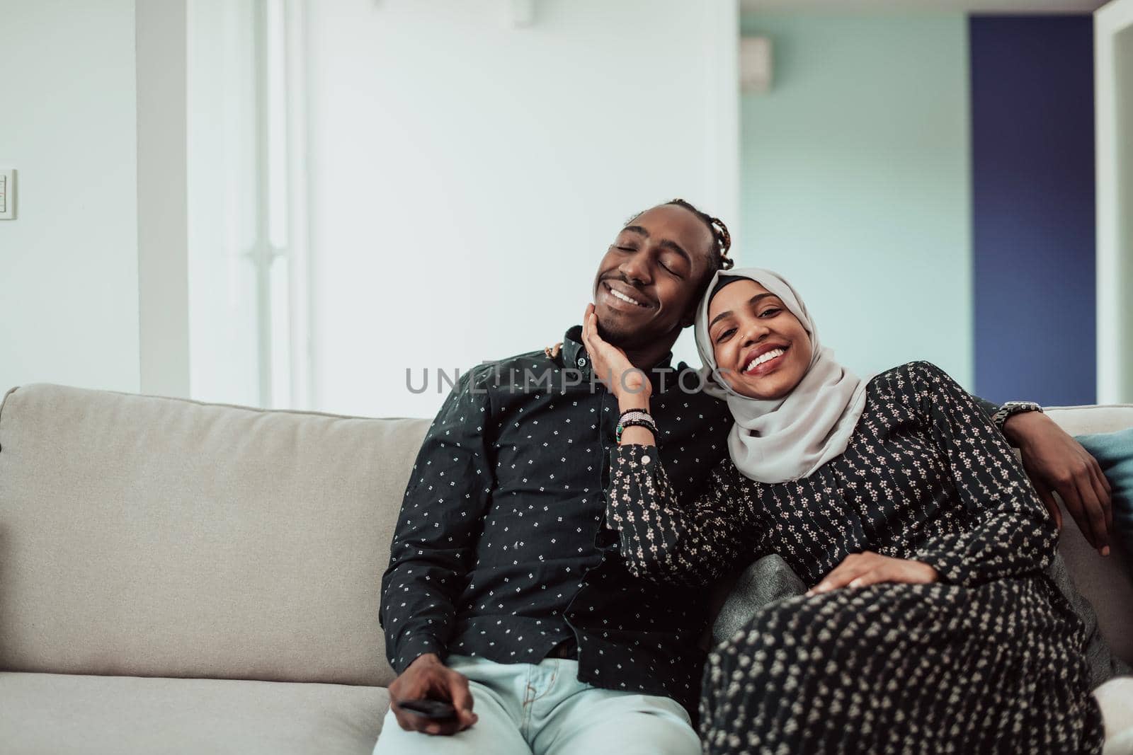 African Couple Sitting On Sofa Watching TV Together. Women Wearing Islamic Hijab Clothes. High-quality photo