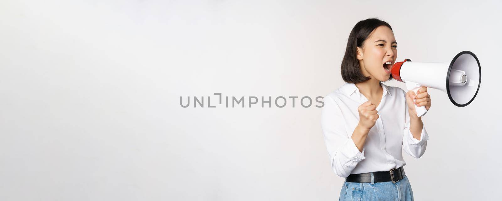 Enthusiastic asian woman, girl activist shouting at protest, using megaphone, looking confident, talking in loudspeaker, protesting, standing over white background.