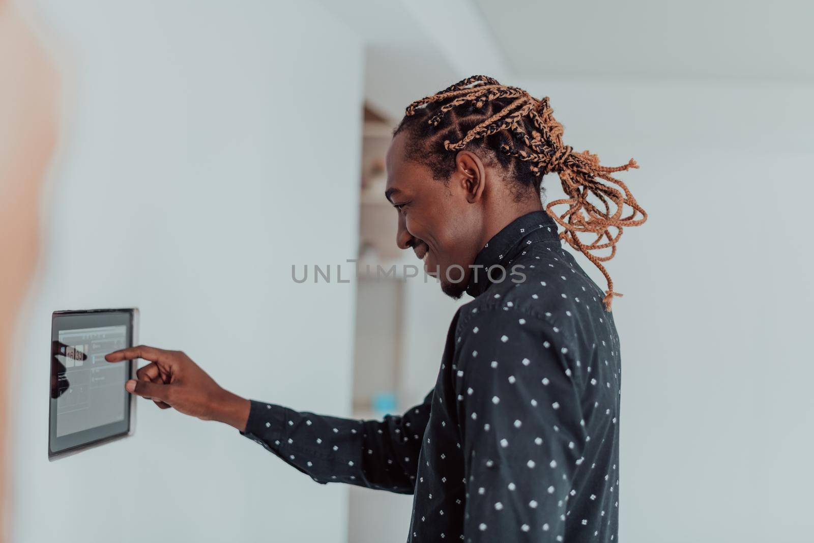 Smiling African American man using modern smart home system, controller on wall, positive young man switching temperature on thermostat or activating security alarm in apartment. High quality photo