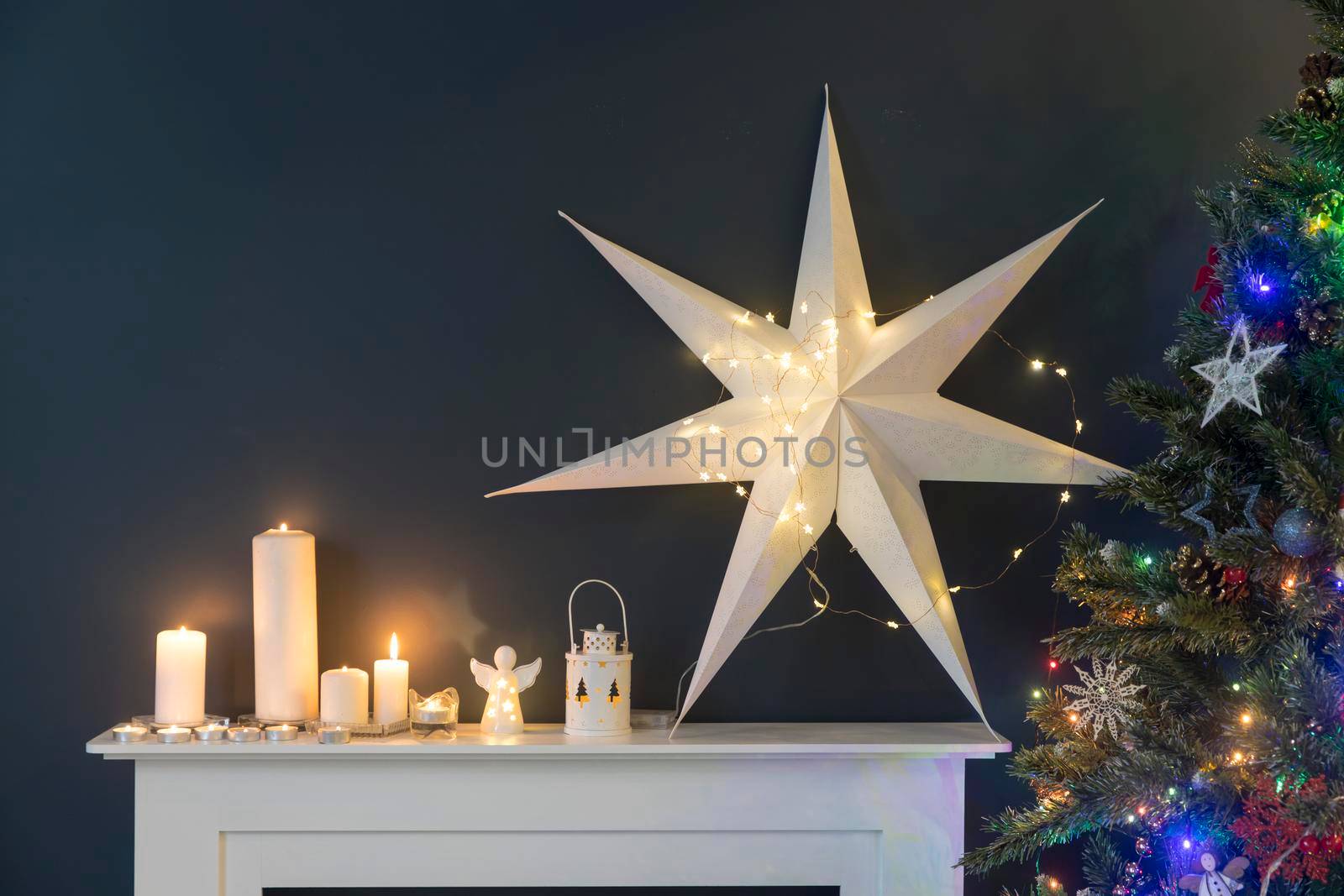 Home decoration before Christmas. Decorated Christmas tree with garland lights, rocking horse, gifts wrapped in elegant paper, artificial fireplace with a large white paper star and candles.