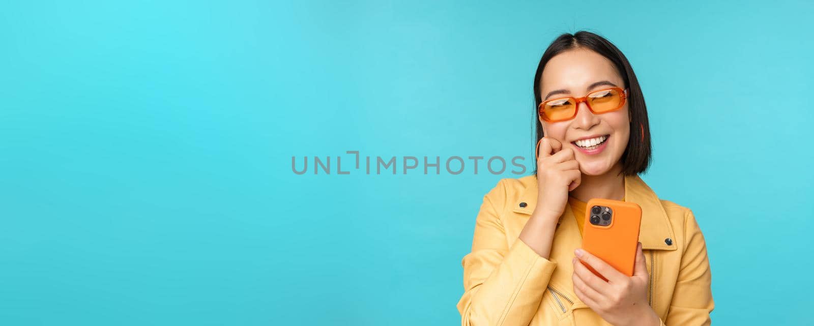 Happy stylish asian girl using smartphone and laughing, smiling at camera, standing over blue background. Copy space