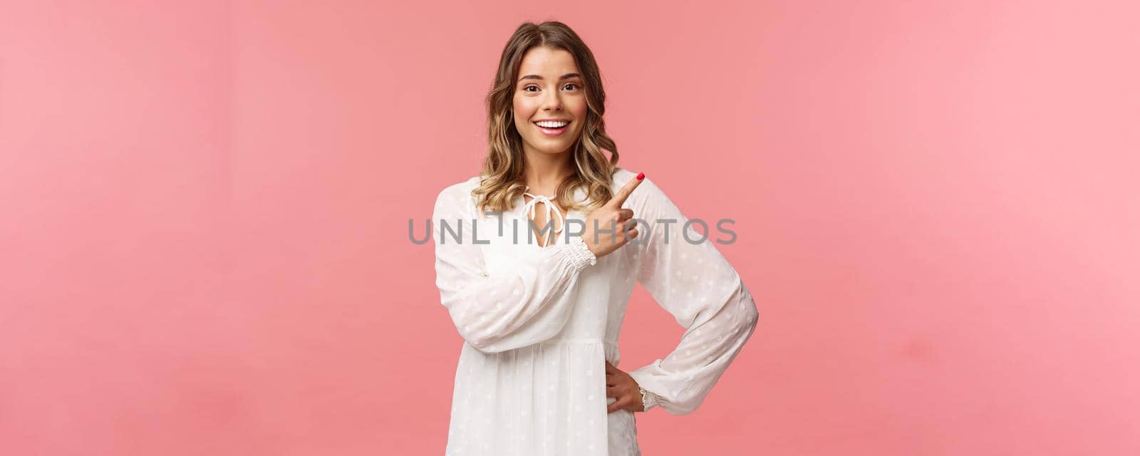 Upbeat good-looking caucasian blond girl in white dress, pointing upper right corner and smiling at camera with interested, curious face discuss special promo spring discounts, pink background by Benzoix