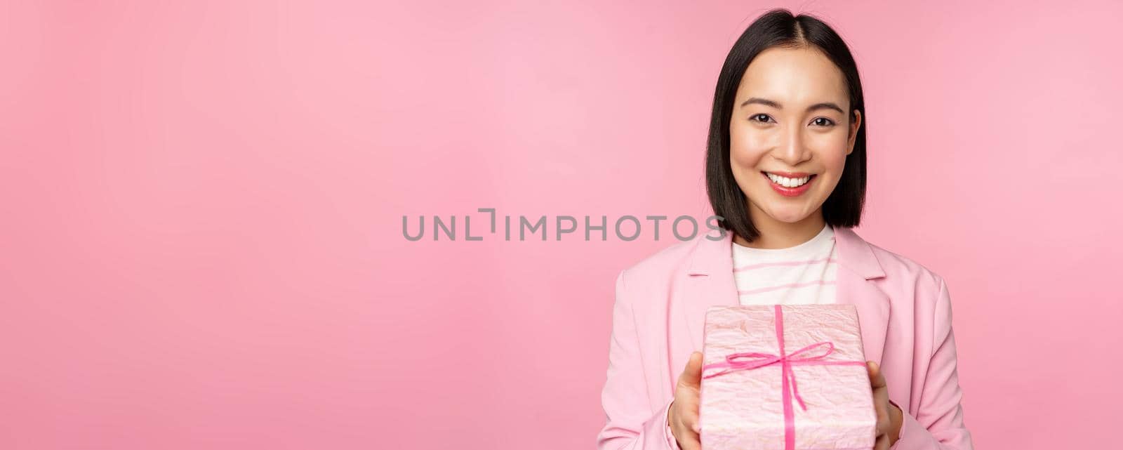 Smiling asian businesswoman in suit, giving you gift in wrapped box, standing over pink background by Benzoix