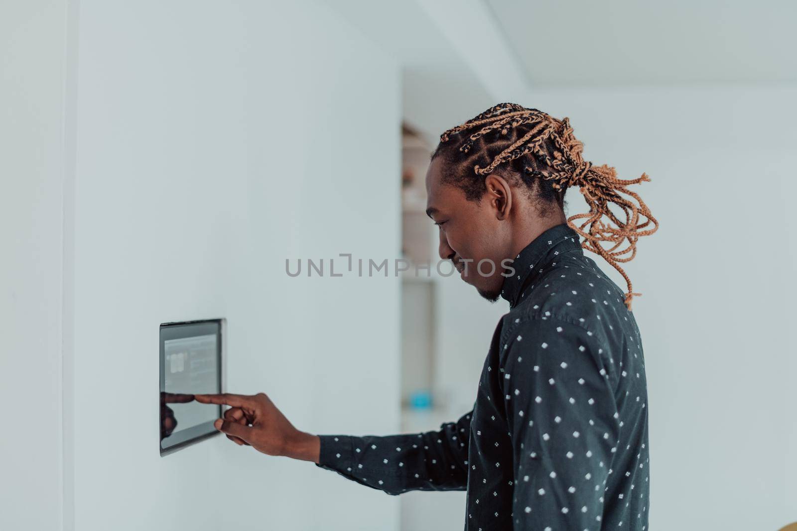Smiling African American man using modern smart home system, controller on wall, positive young man switching temperature on thermostat or activating security alarm in apartment by dotshock