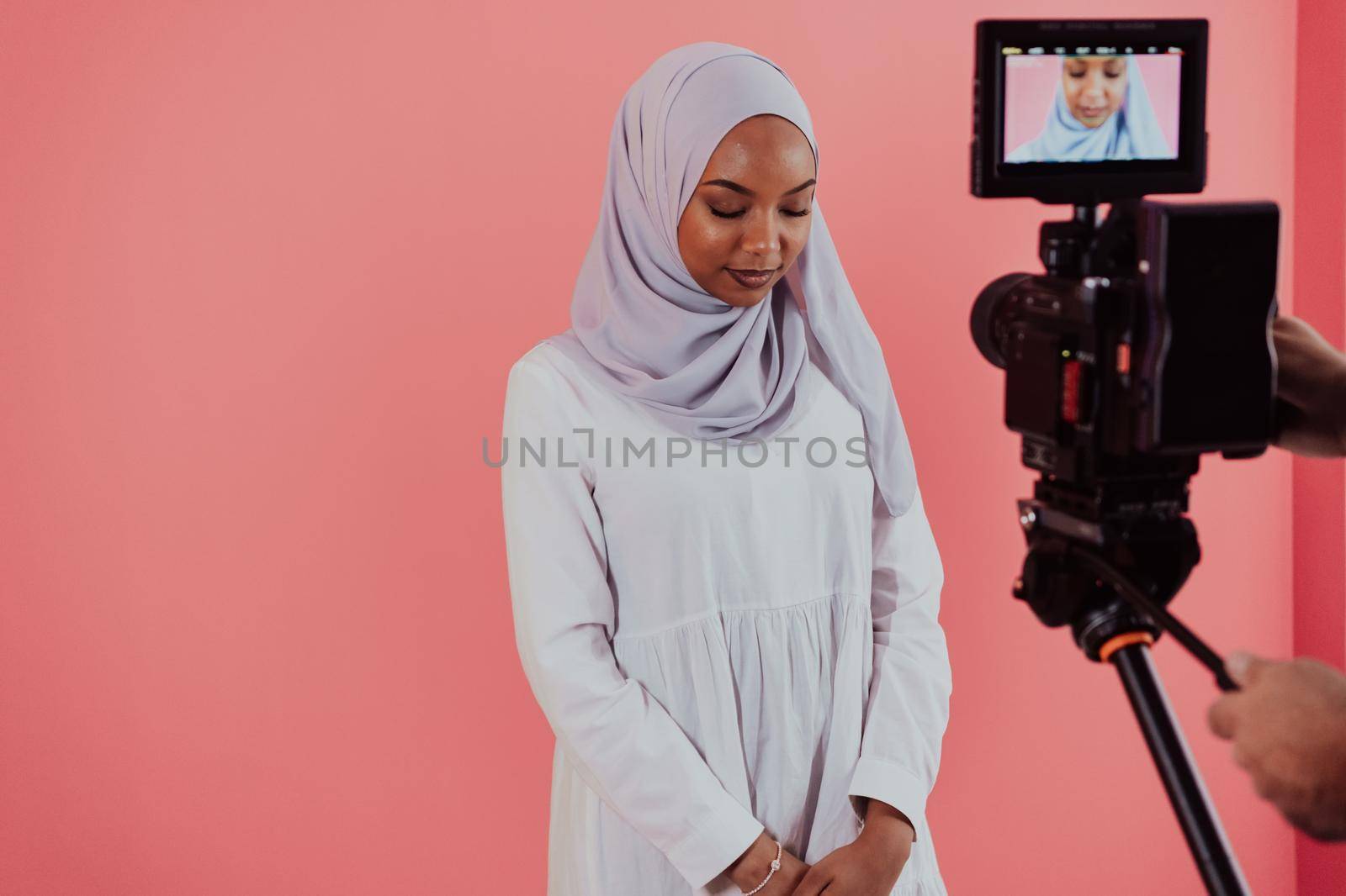 Videographer in digital studio recording video on professional camera by shooting female Muslim woman wearing hijab scarf plastic pink background. High-quality photo