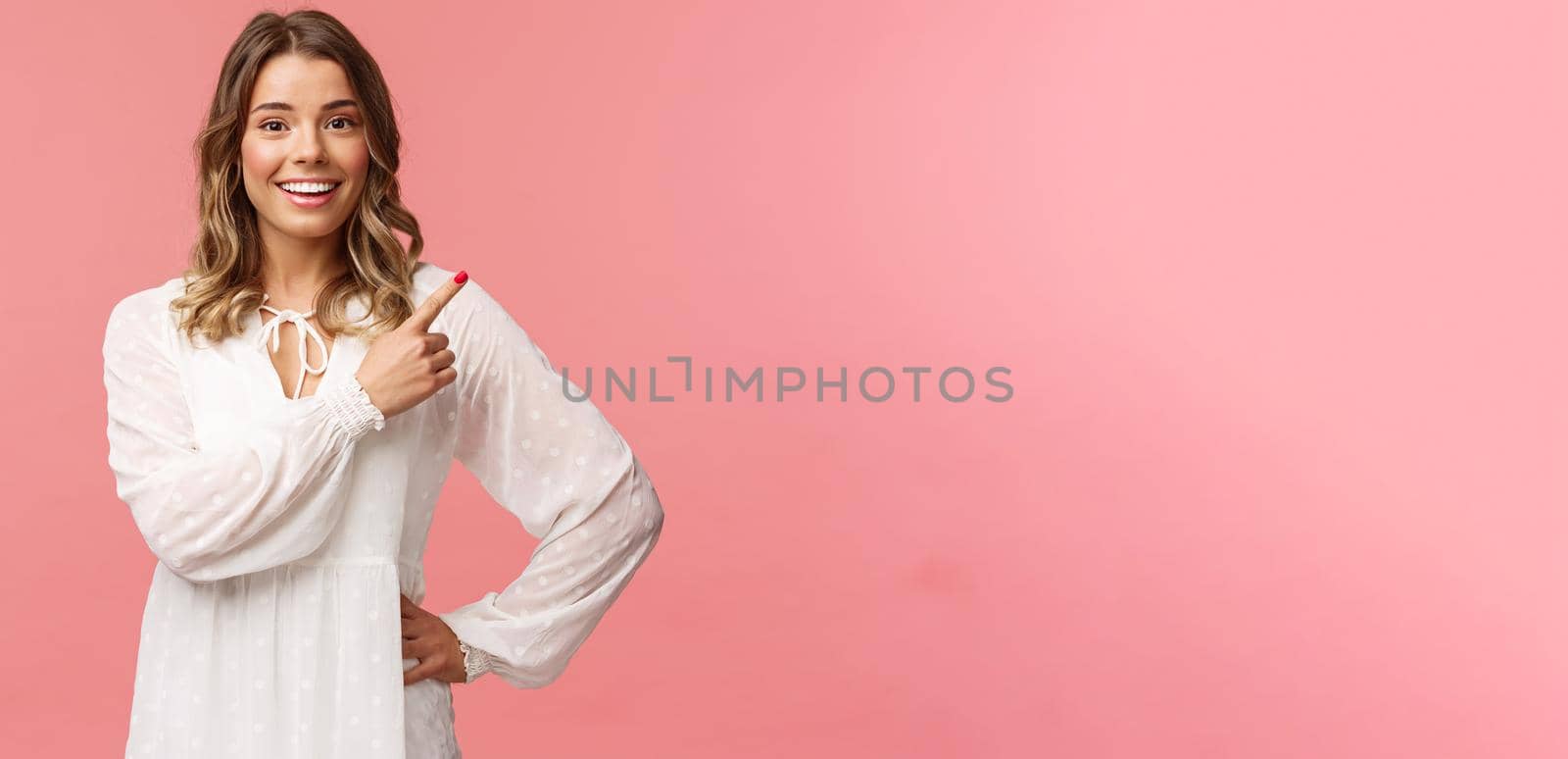 Upbeat good-looking caucasian blond girl in white dress, pointing upper right corner and smiling at camera with interested, curious face discuss special promo spring discounts, pink background.