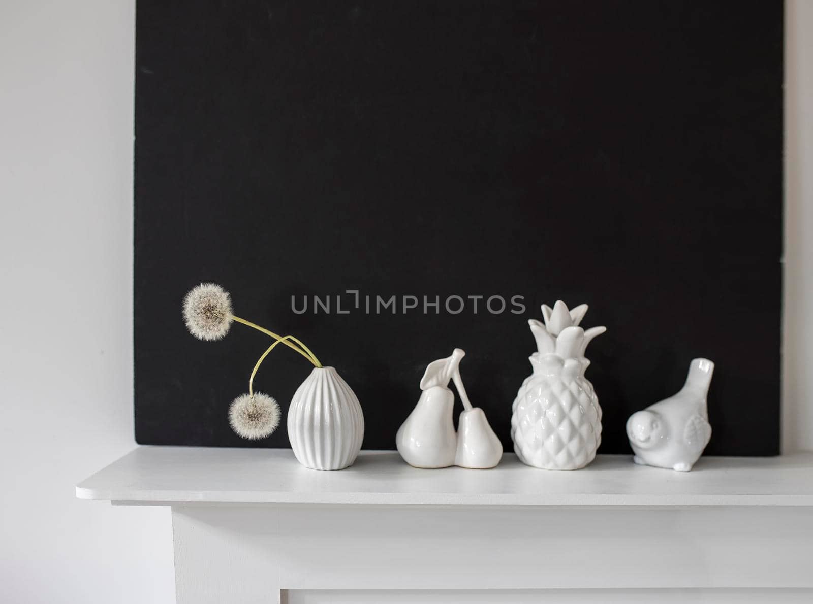 White figurines of pineapple, pear, bird, a vase with two dandelions on the background of a black painting on a white table. Space for text