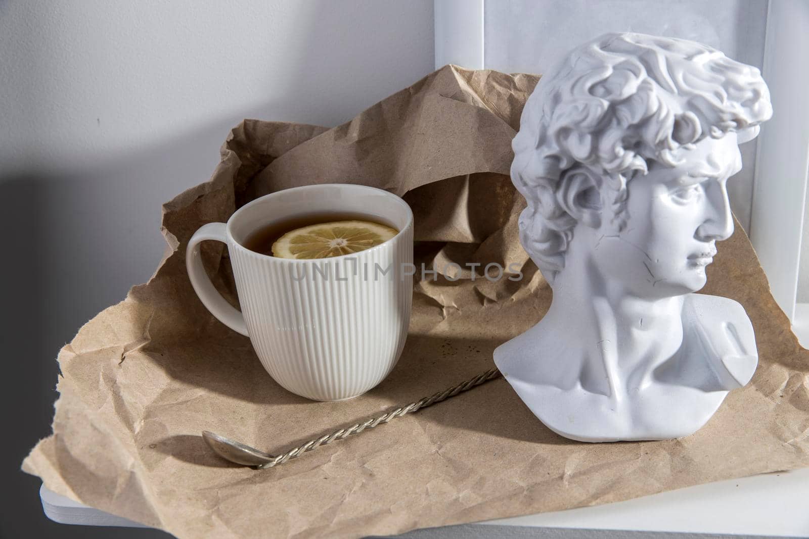 A white corrugated cup with tea and lemon, with a plaster head sculpture of Apollo and a cupronickel teaspoon on a long handle is on brown craft paper on a wooden table.